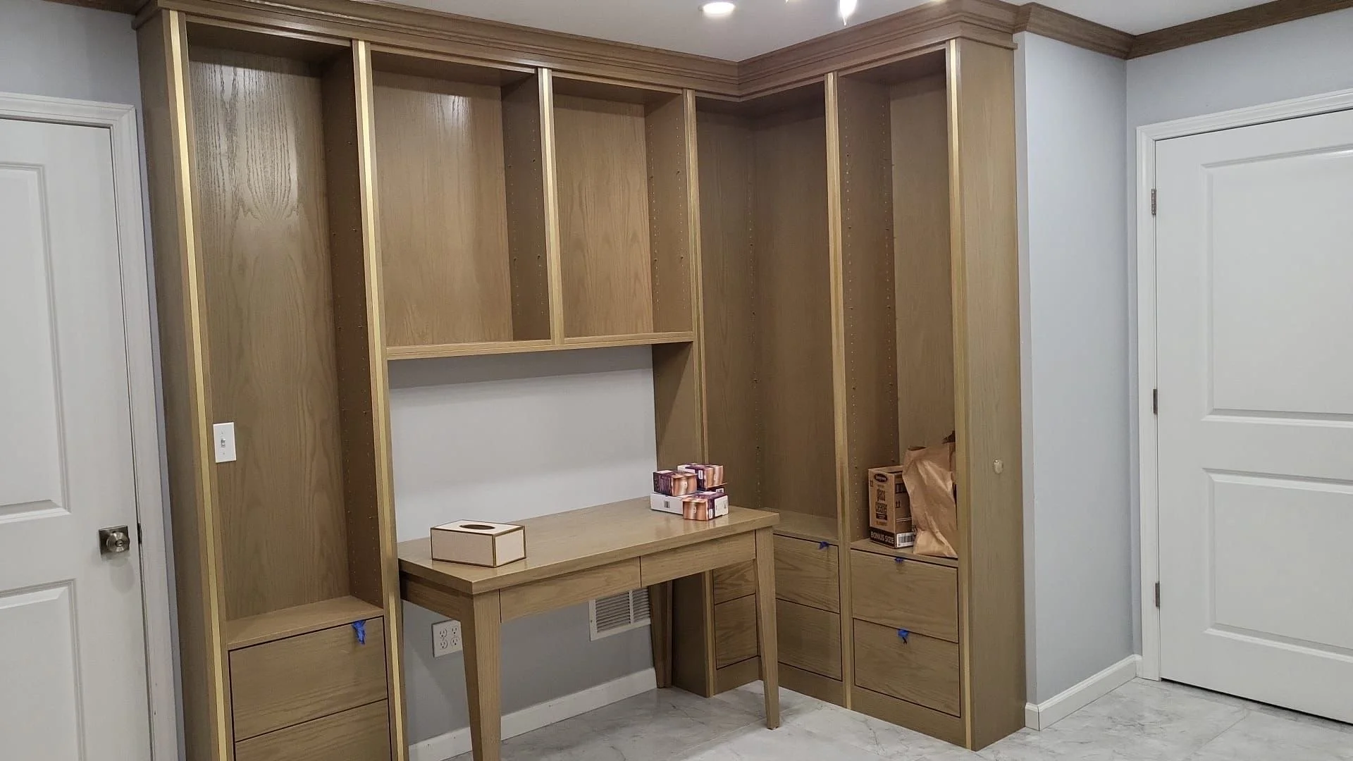 Empty wooden kitchen cabinetry install with a small desk and box of painted wood samples in a room with white doors and gray walls.