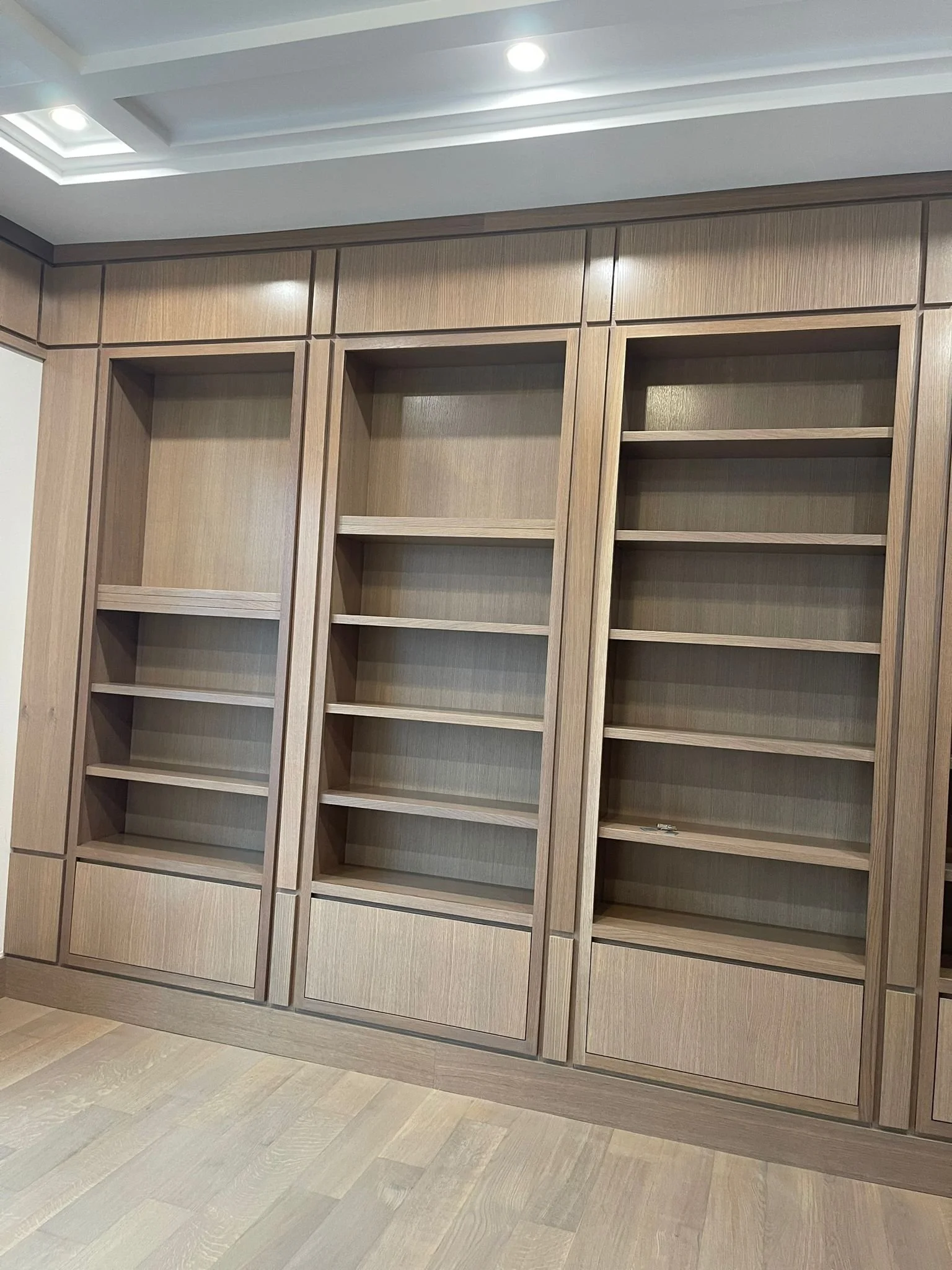 Empty wooden built-in bookshelves with no books in a room with light-colored wood flooring and ceiling lights.