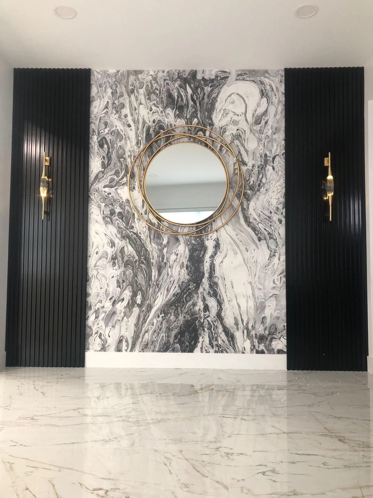 Decorative wall with black and white marble design, large circular mirror with gold rings, flanked by black ribbed wall panels with gold sconces, marble floor.