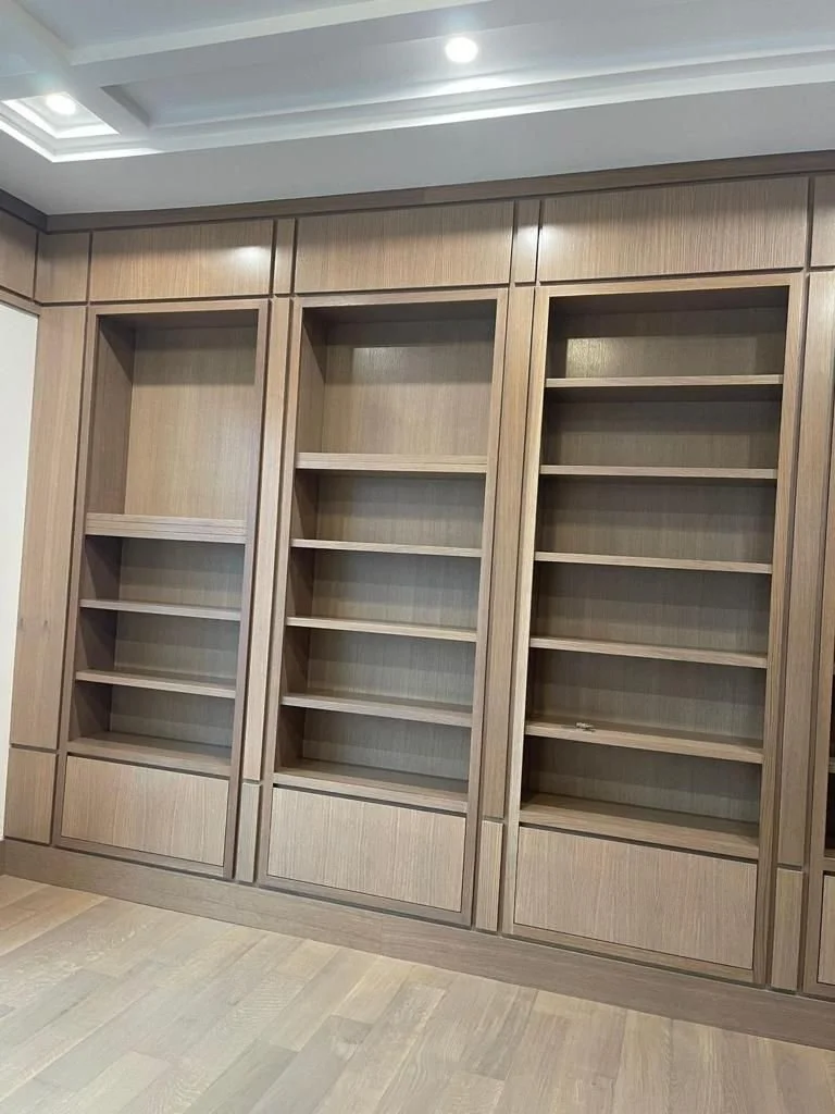 Empty built-in wooden bookshelf with multiple shelves against a wooden-paneled wall in a room with light-colored hardwood flooring.