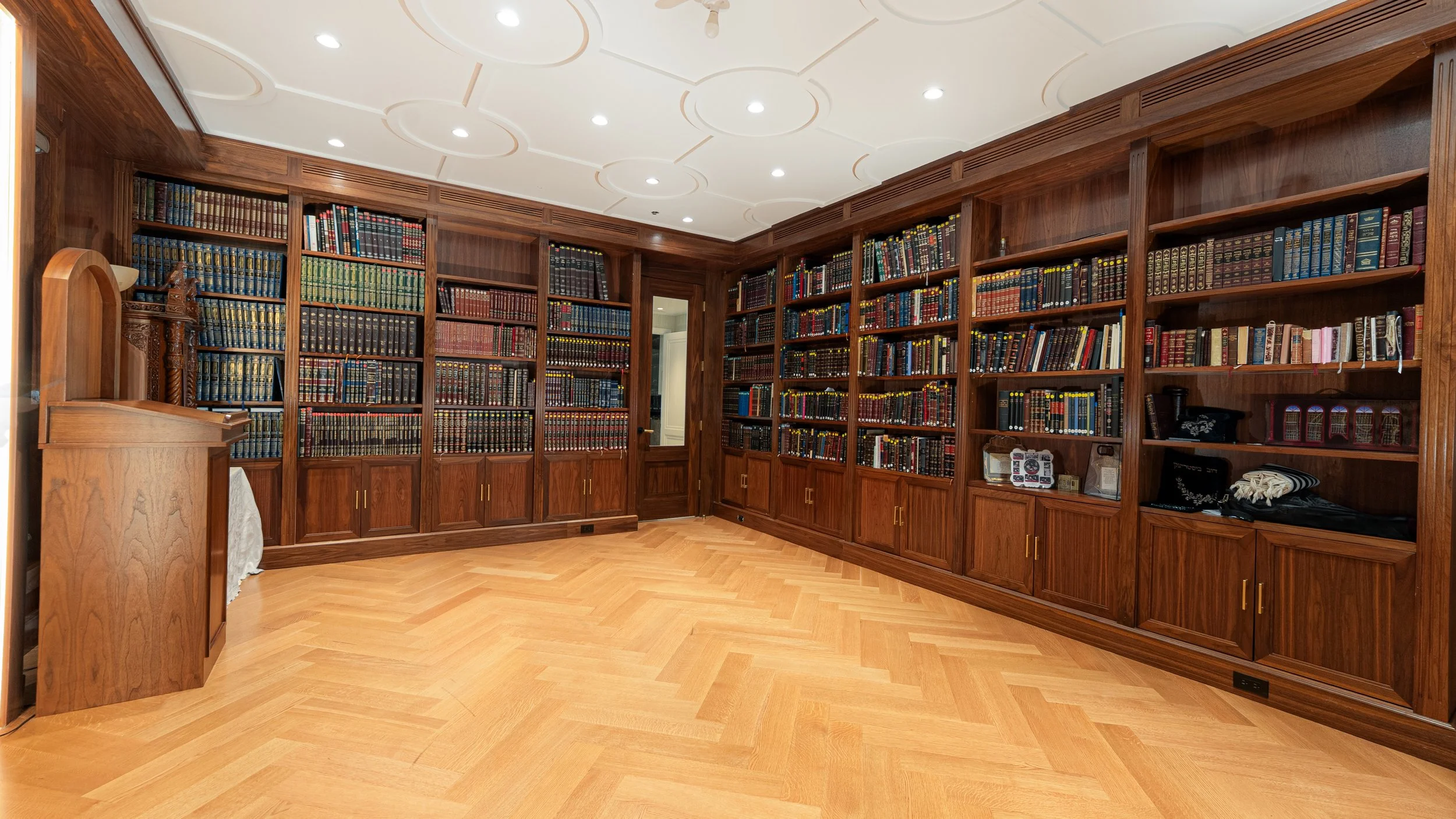 A spacious home library with wooden bookshelves filled with numerous books, a hardwood floor, and a white ceiling with recessed lighting.