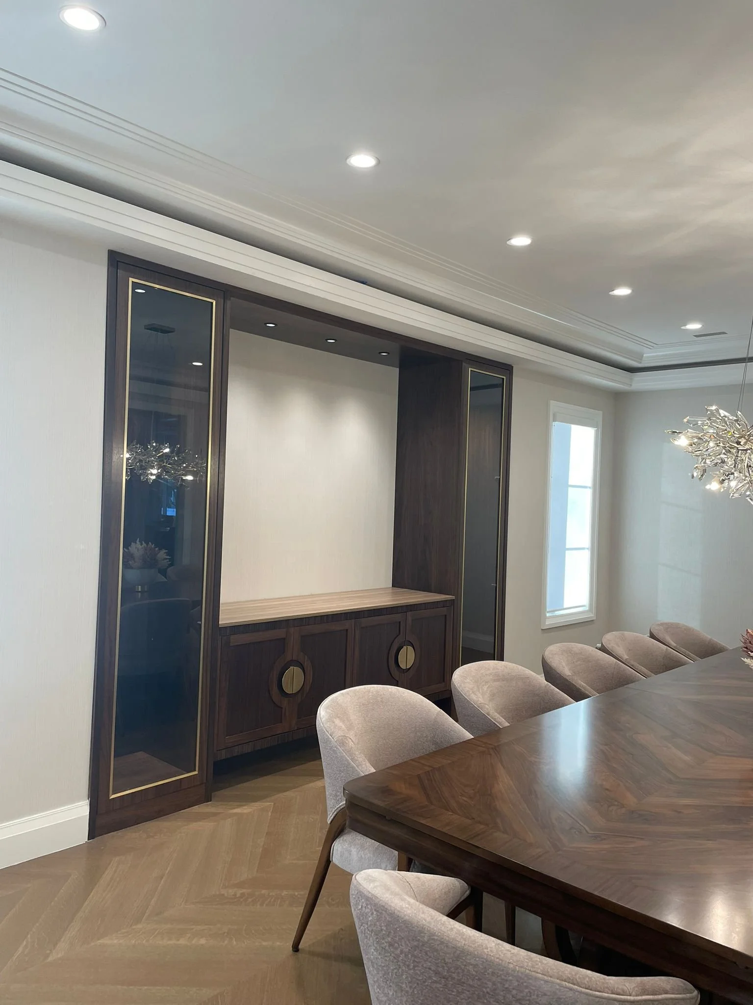 A modern dining room with a wooden table, beige upholstered chairs, and a built-in dark wood cabinet with glass doors and gold accents. A window is present on the right wall, and a contemporary chandelier hangs from the ceiling.
