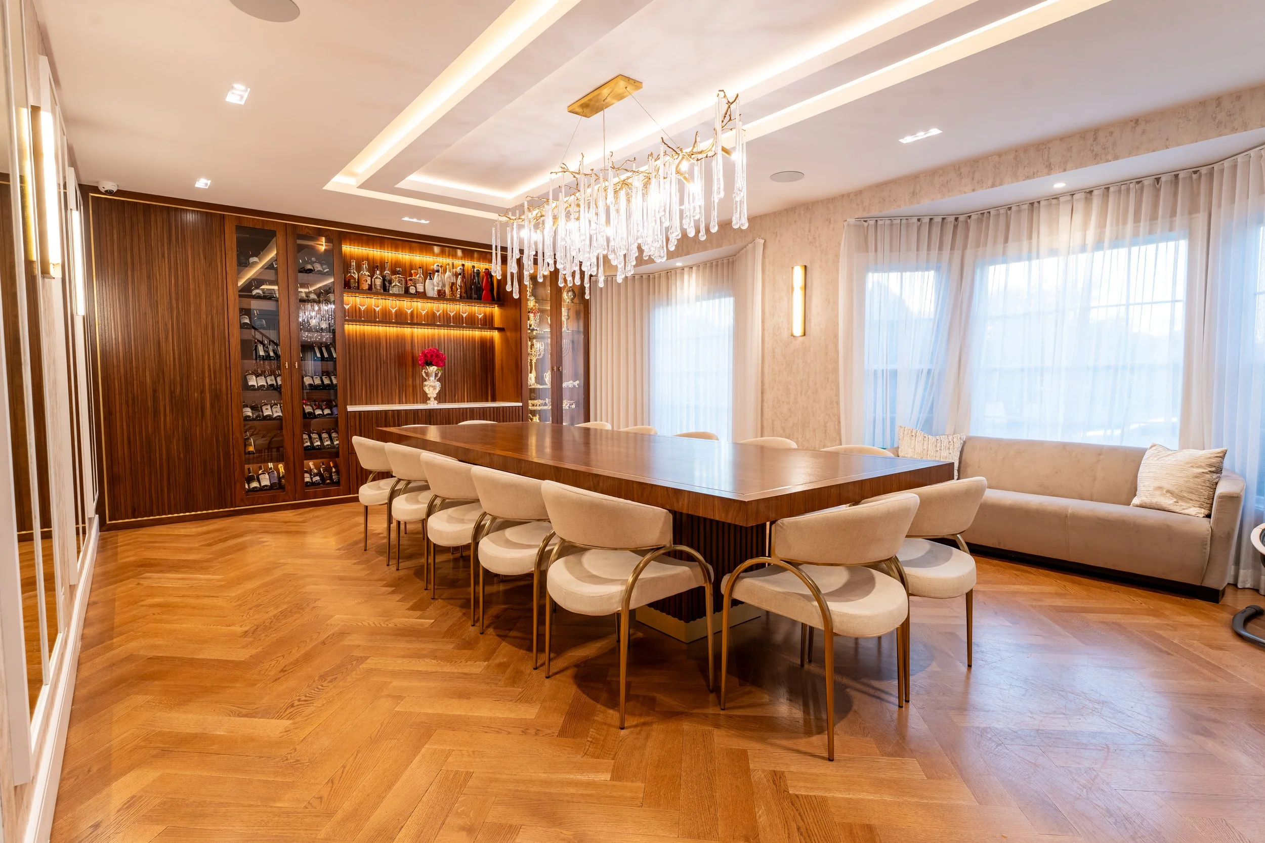 A modern dining room with a large wooden table surrounded by beige upholstered chairs with gold legs. There is a built-in wooden wine cabinet in the back with glass doors, stocked with wine bottles. The room has hardwood floors, large windows with sh