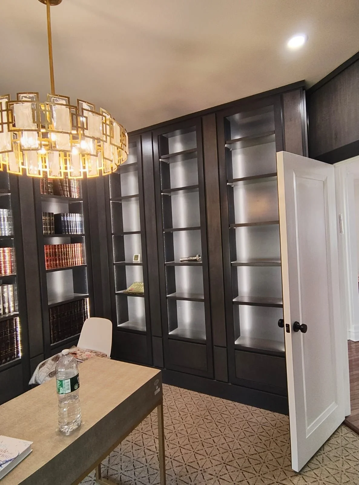 Empty built-in black bookshelves in a room with a patterned beige carpet, a white door, a wooden table with a water bottle, and a white chair.