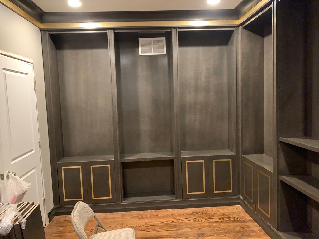 Empty dark gray built-in closet with gold accents on lower cabinets in a room with hardwood floor and part of a white chair in front.