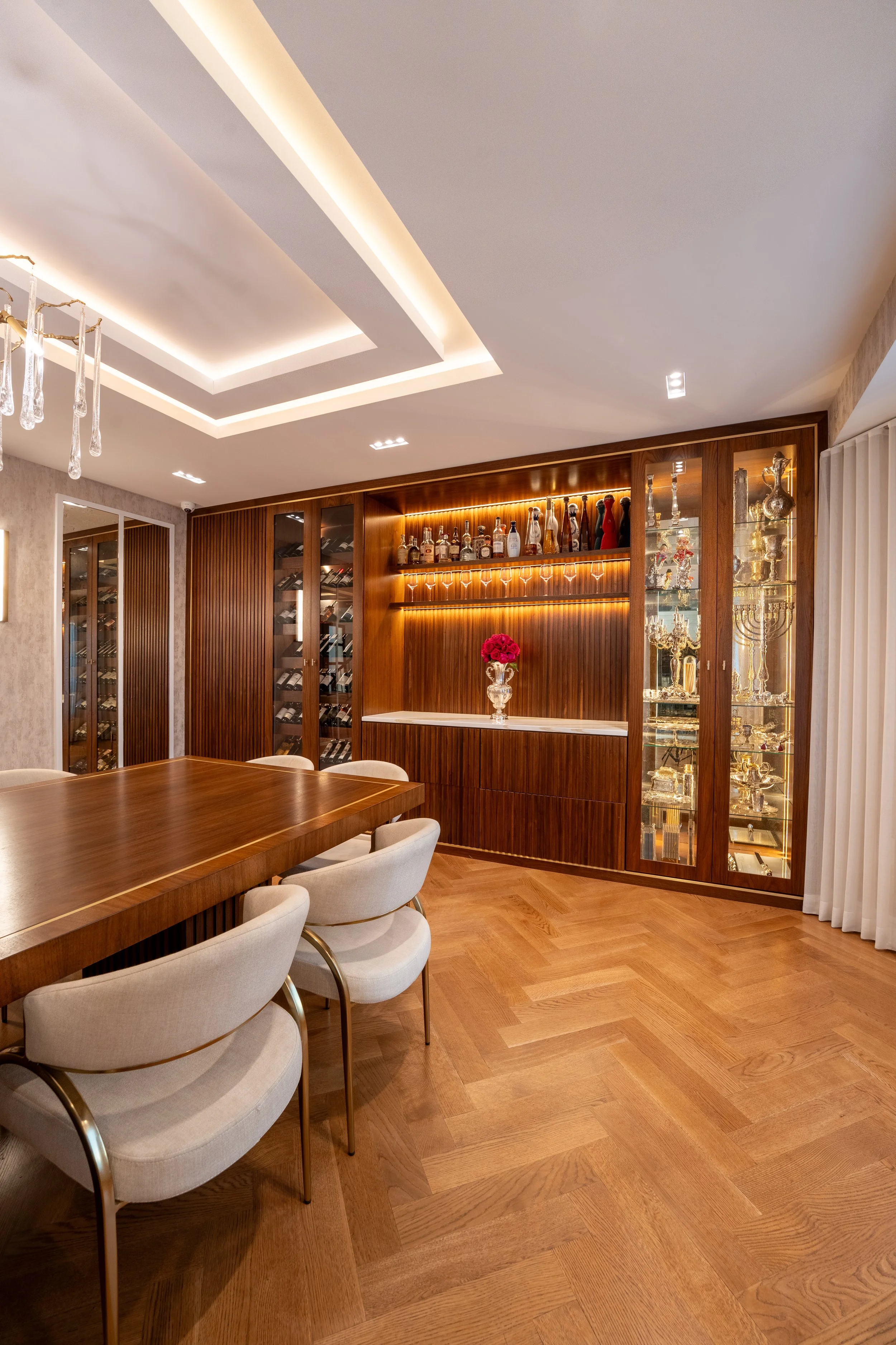Elegant dining room with wooden furniture, a glass display cabinet filled with glassware and collectibles, a wine display on the wall, and white upholstered chairs around a wooden table.