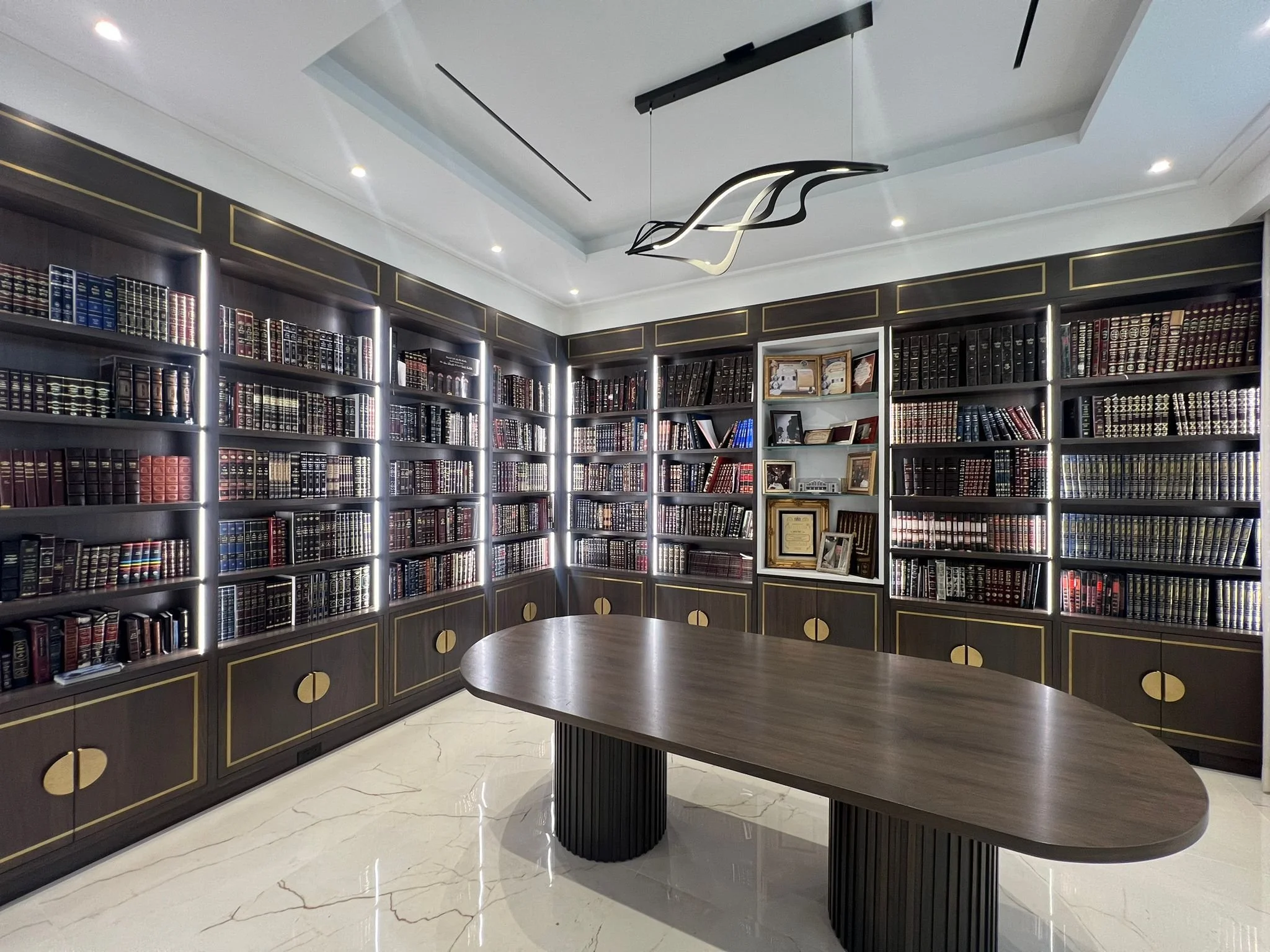 A modern, well-lit library with dark wood bookshelves containing numerous books, a large oval wooden table in the center, and a contemporary ceiling light fixture.