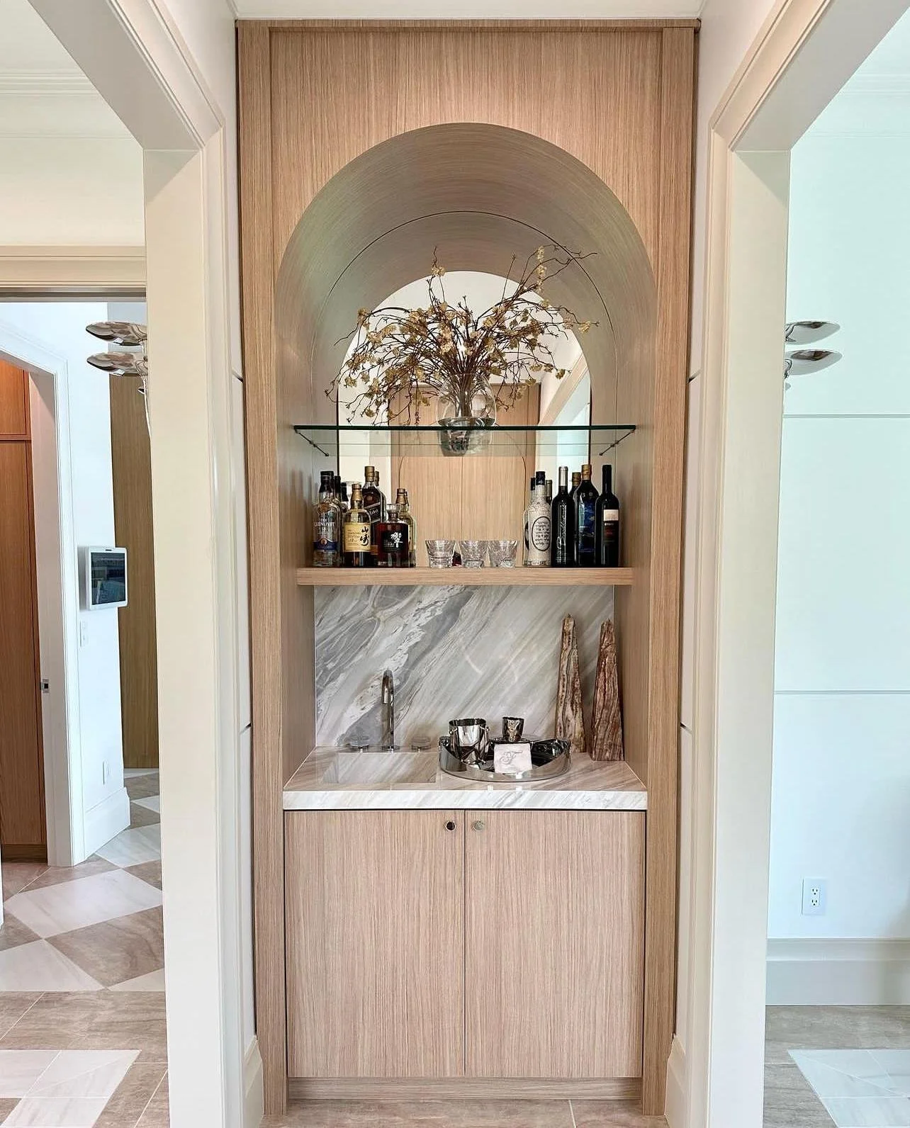 Built-in cabinet with a marble backsplash, glass shelf with decorative glassware, and a collection of liquor bottles and a vase with dried flowers on top.