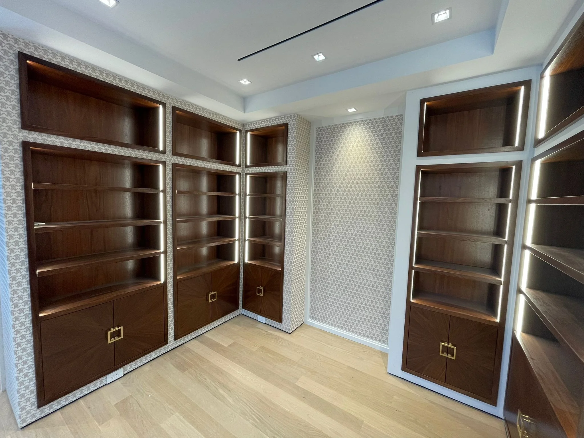 A corner of a room with wooden open shelves and cabinets, patterned wallpaper on the walls, and recessed ceiling lights.