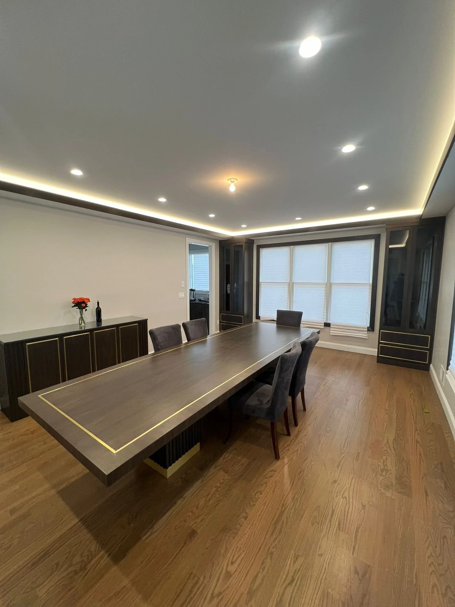 A modern dining room with a large dark wood table, six gray upholstered chairs, a sideboard with a vase and wine bottle, a large window with blinds, and recessed ceiling lights with some LED strip lighting.