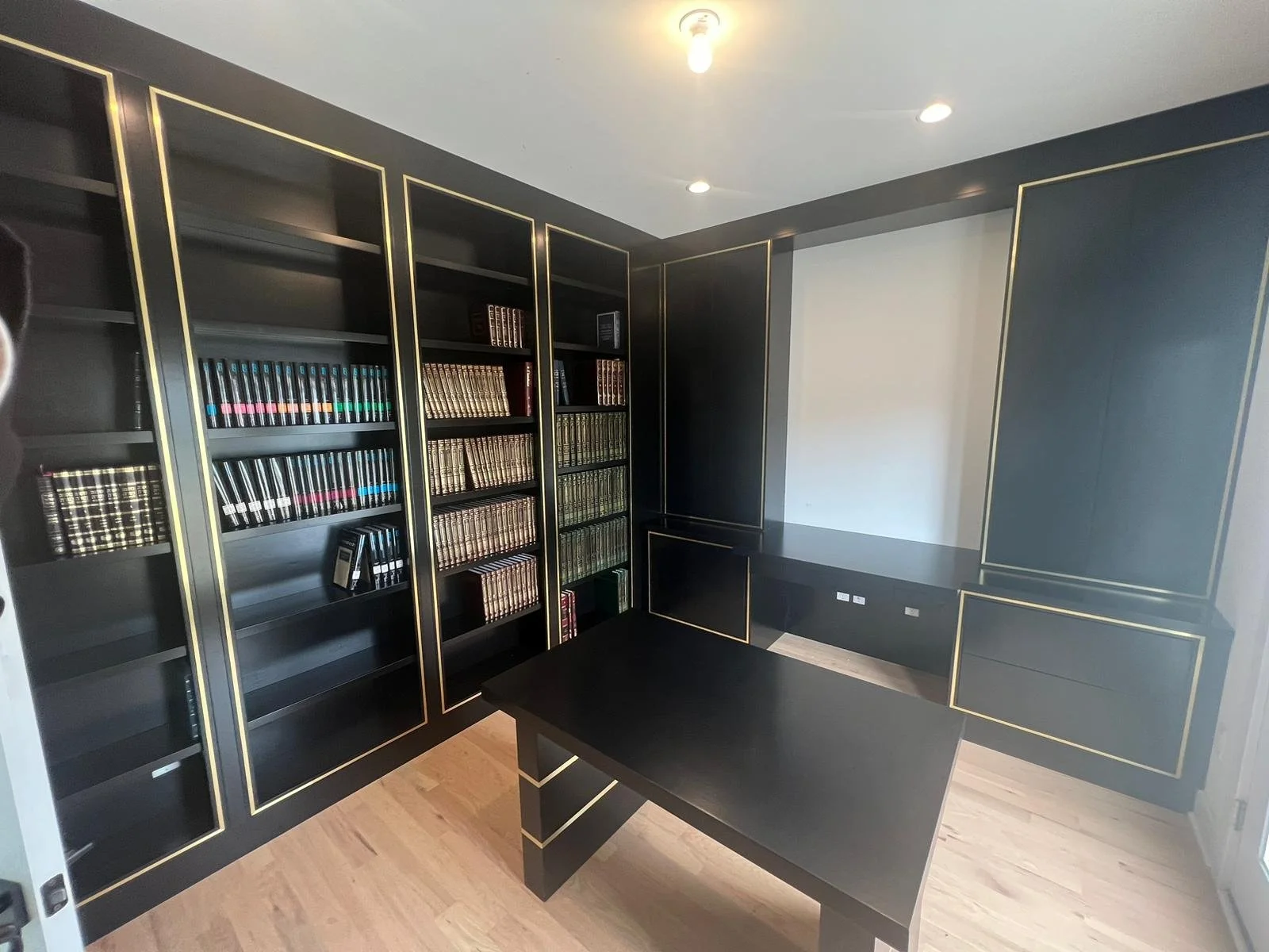 Black built-in bookshelf and cabinet with gold trim, filled with colorful books, in a modern office room with light wooden flooring, and a black desk in the foreground.
