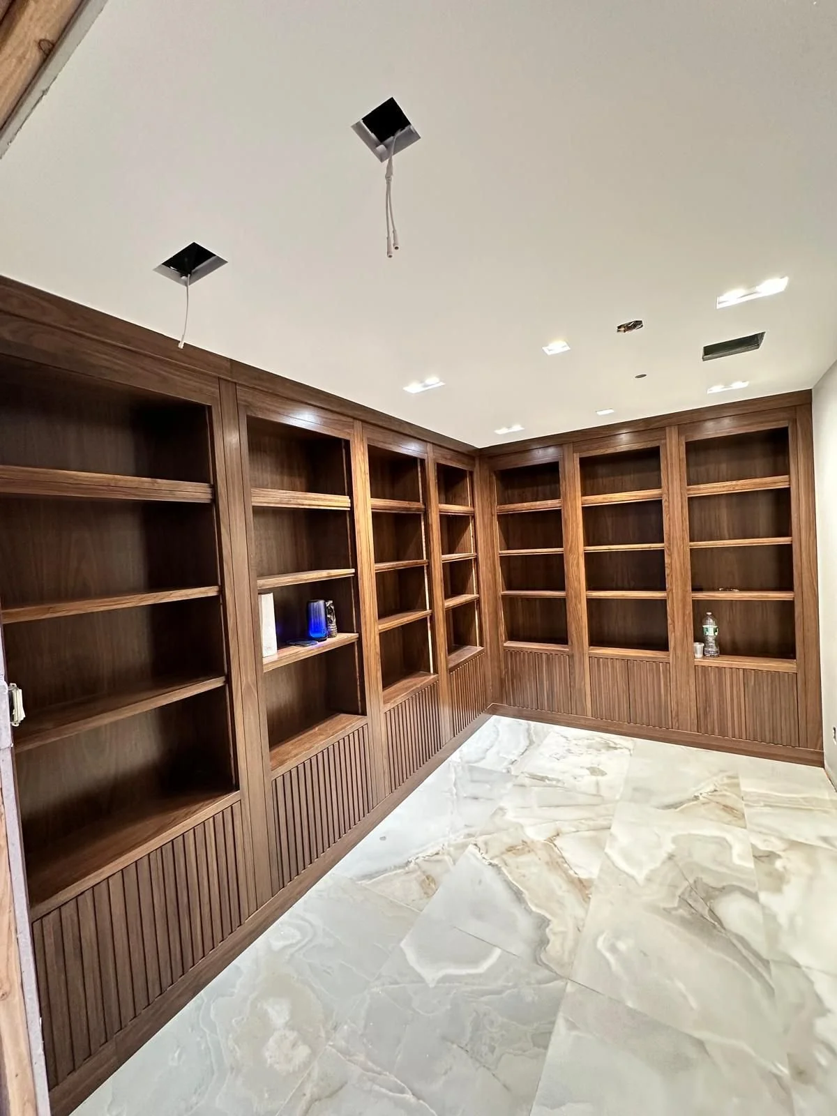 Empty wooden bookshelves in a room under construction with a marble floor and a white ceiling with incomplete lighting fixtures.