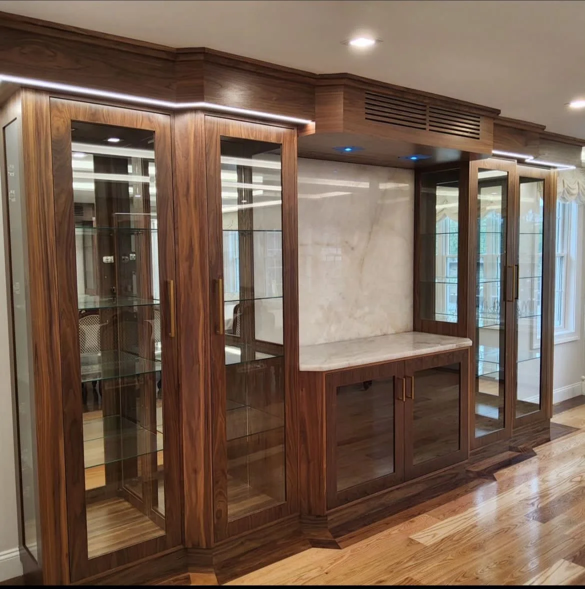 A custom-built wooden display cabinet with glass doors and shelves, featured in a room with wooden flooring and a marble wall backdrop.