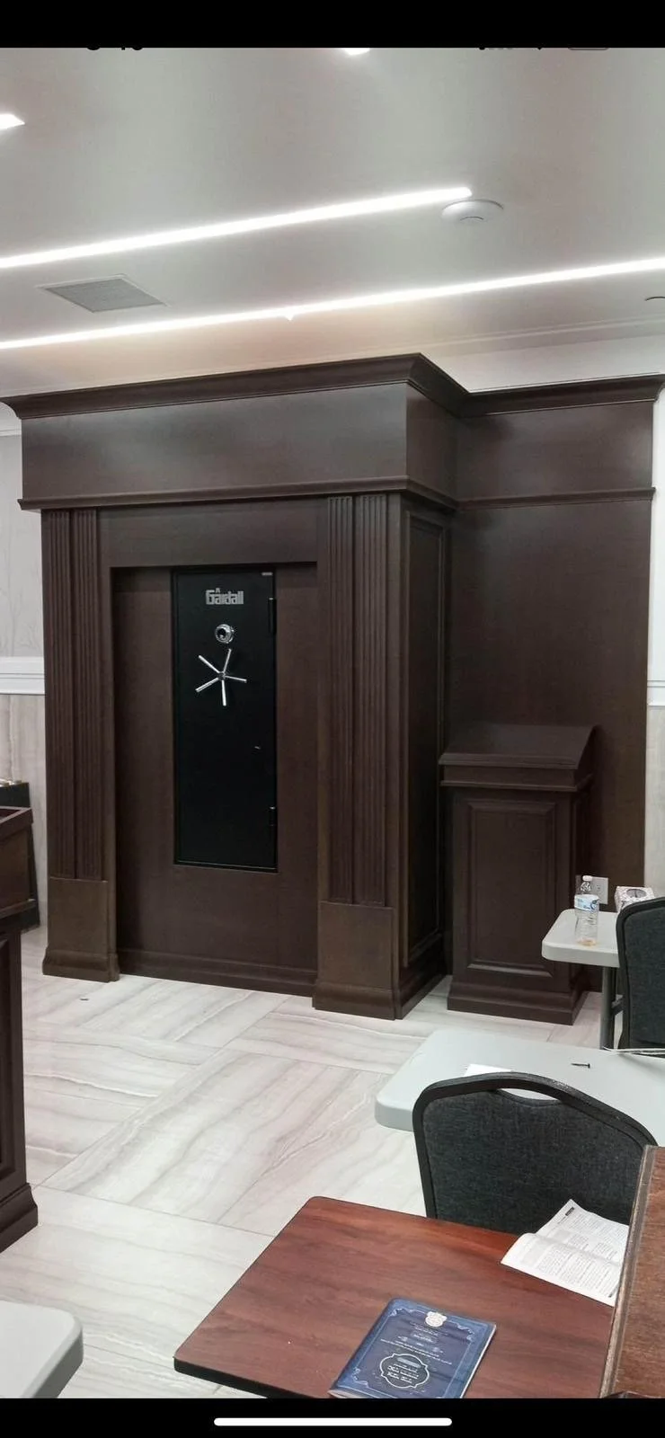 A dark wooden wall safe with a clock on it, situated in a room with a wooden corner piece and a table with papers, water bottles, and chairs.