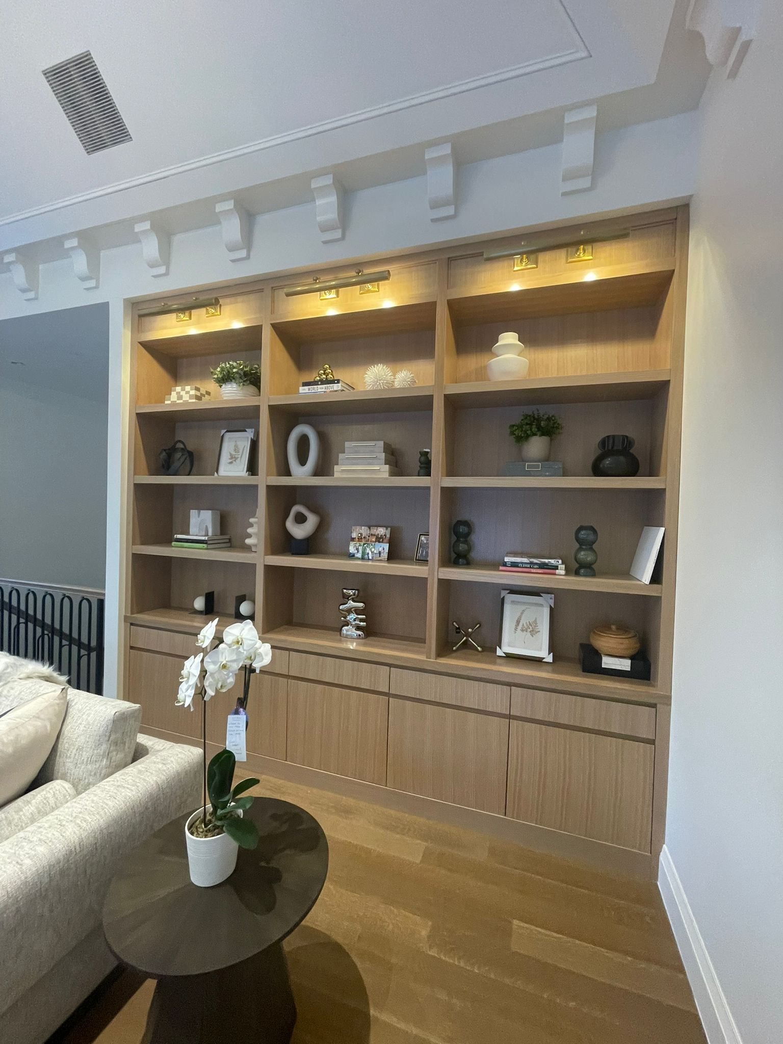 Wooden wall unit with decorative objects and books, located in a living room with a beige couch and a small round table with a potted orchid.