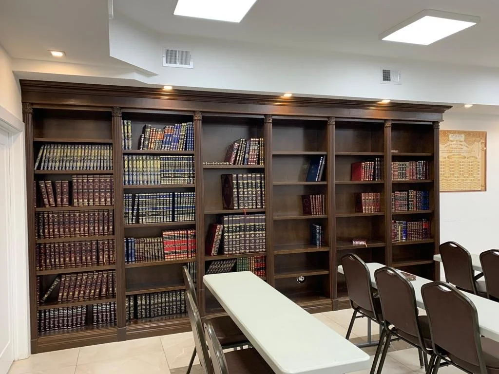 A large wooden bookshelf filled with books, located in a well-lit room with white walls and multiple tables and chairs.