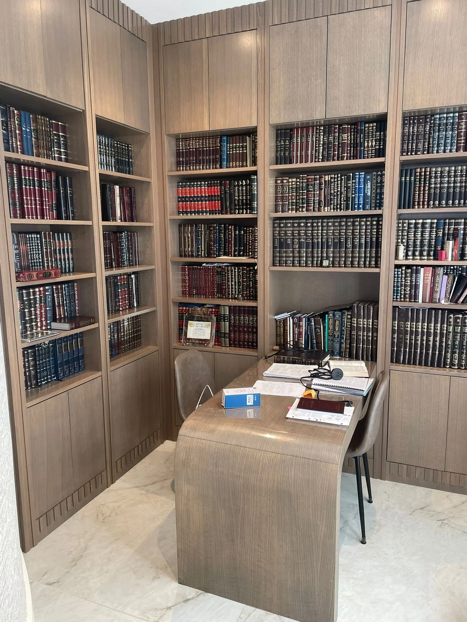 A small library or study room with built-in wooden bookshelves filled with numerous books and a wooden desk with papers, a closed notebook, a box, and a headphones resting on it, and two chairs.