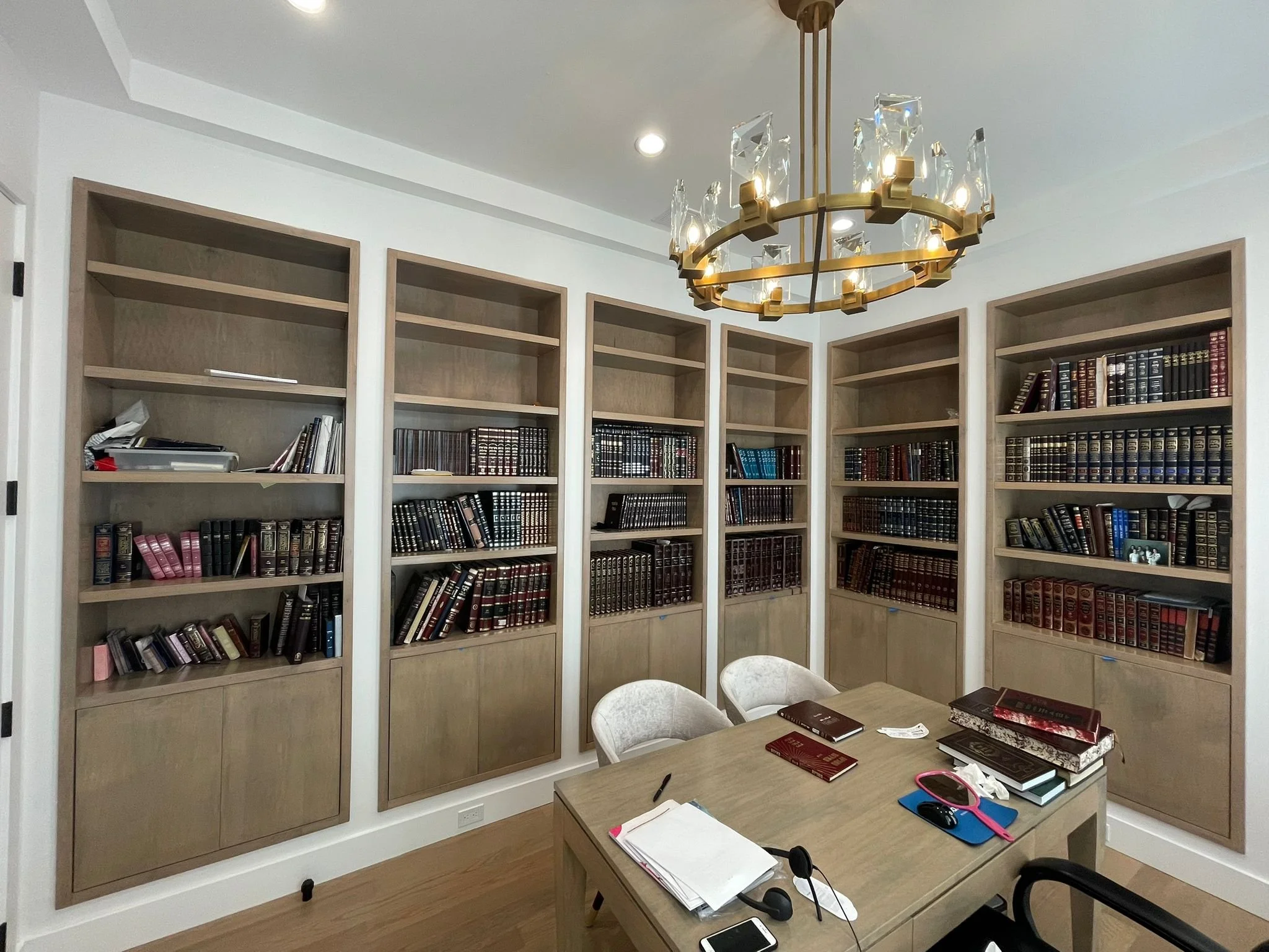 A home office with a large, filled wooden bookshelf, a wooden table with books, papers, a computer mouse, a pink headband, a smartphone, and a pair of headphones, and a chandelier hanging from the ceiling.