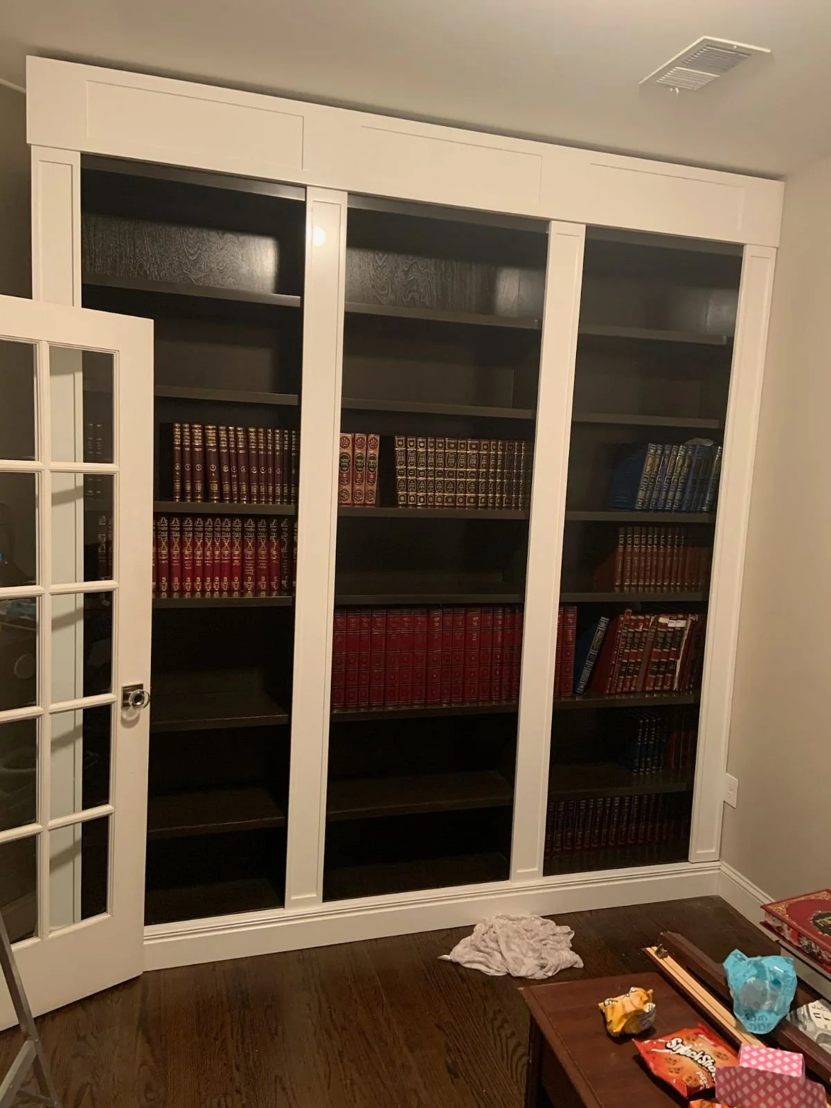 Empty bookshelf with dark wood interior and white trim in a room with a closed glass-paned door, small table with snacks, and a crumpled cloth on the floor.