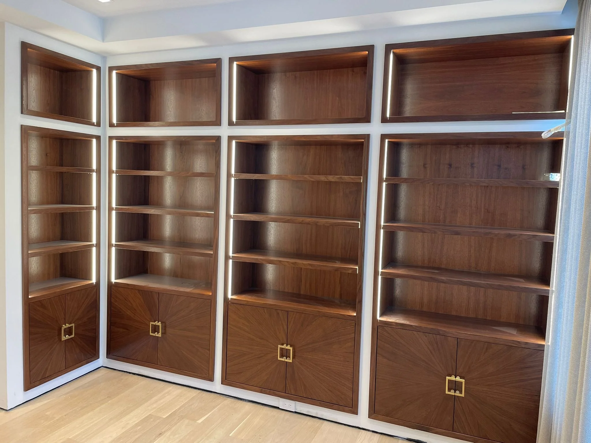 Empty wooden bookshelf with multiple shelves and closed cabinets at the bottom, built into a white wall with lighting accents.