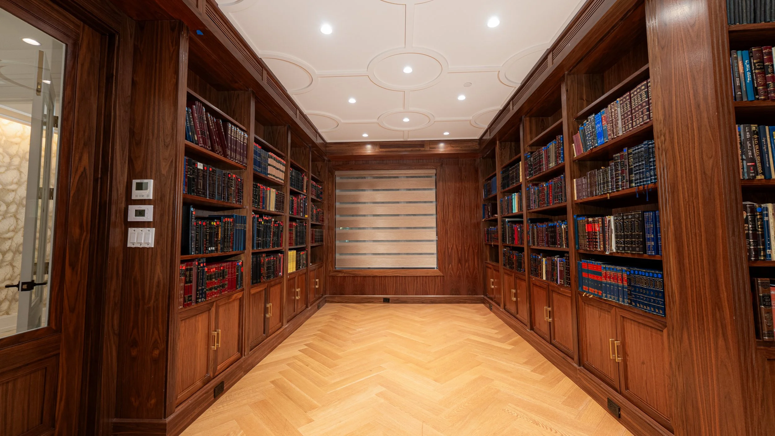 Wood-paneled library with built-in bookshelves filled with books, a window with a beige roller shade, hardwood floor, and ceiling lights.