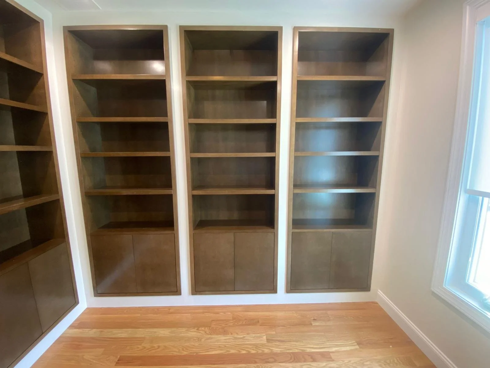 Empty wooden bookshelves with four sections, three with open shelves and lower closed compartments, in a room with a window and hardwood floor.
