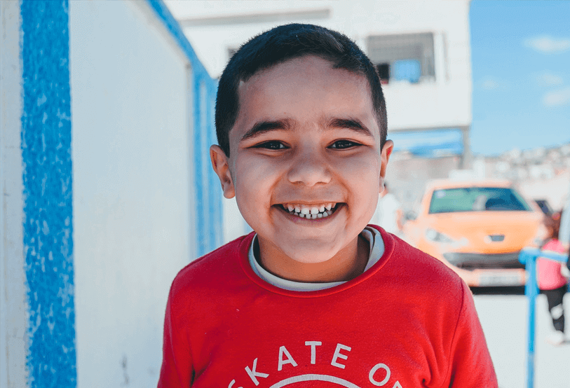 Young boy smiling outdoors, wearing a red shirt with white lettering, in front of a blue and white wall, with cars and people in the background.