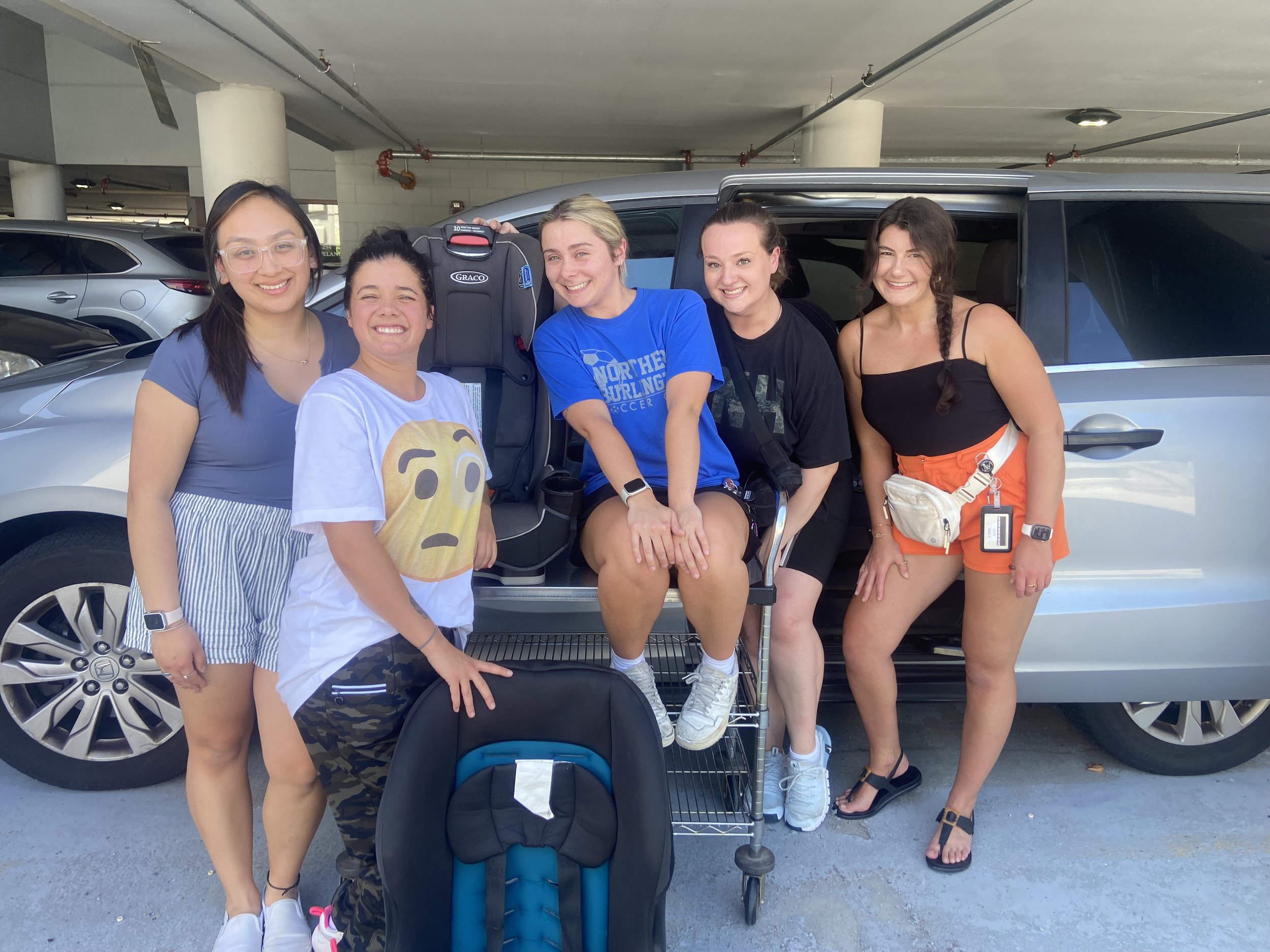 Five young women standing in front of a car in a parking garage, smiling, with a car seat set on a stroller in front of them. They just finished PT1 transportation training!