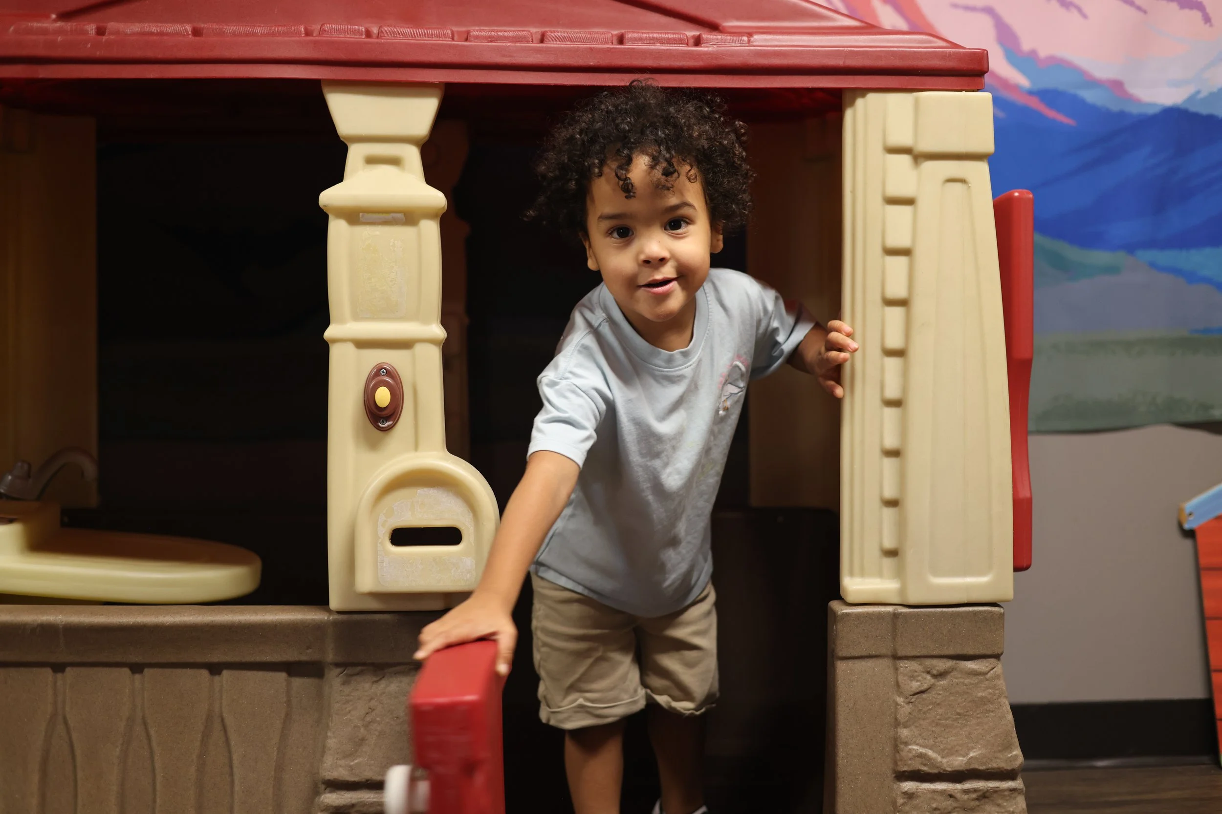 A young boy with curly hair wearing a gray t-shirt and beige shorts inside a plastic playhouse at an indoor play area.