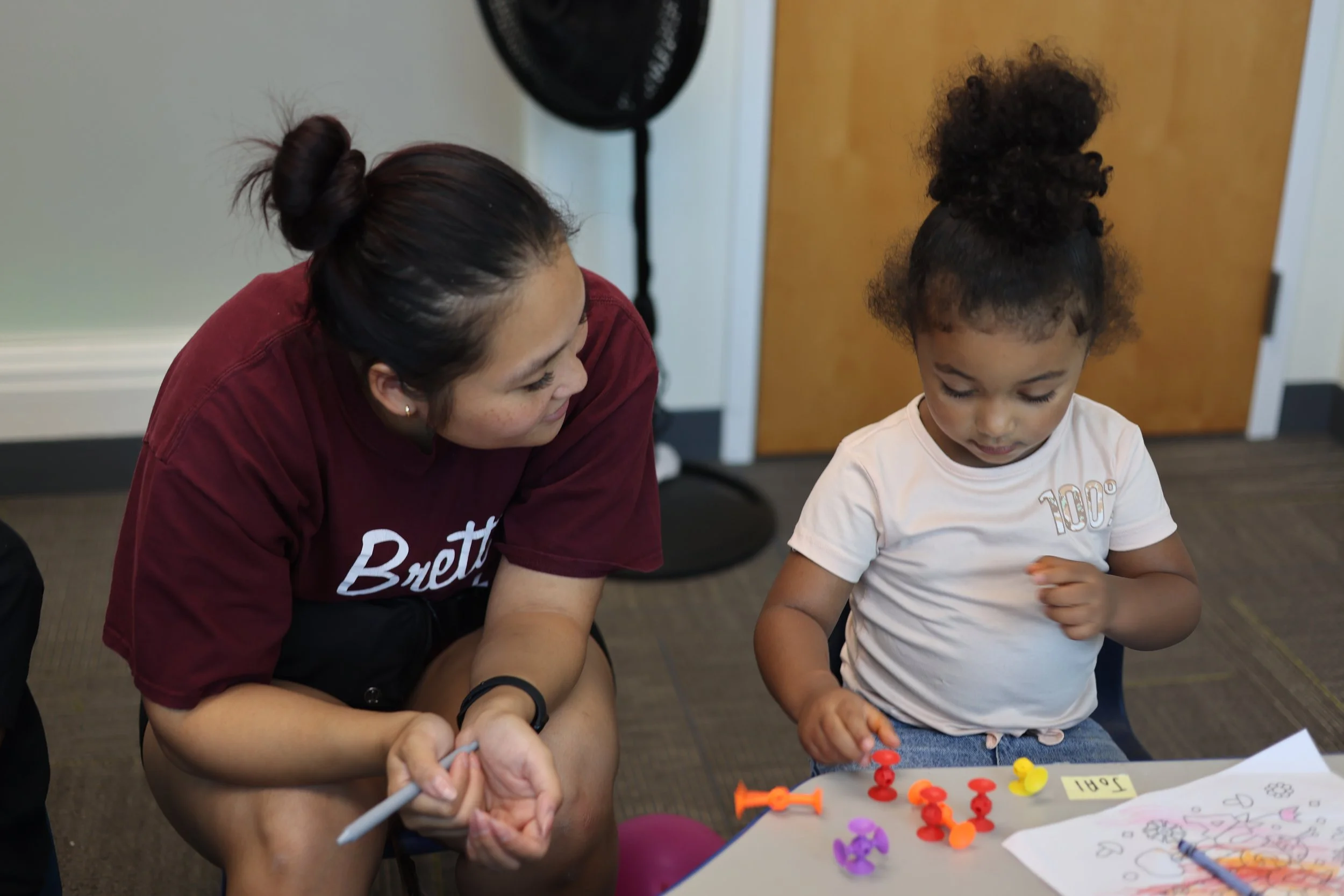 A woman and a young girl engaged in a game or activity at a table, with colorful plastic pushpins and a drawing or coloring sheet, indoors in a room with a door and a fan in the background.