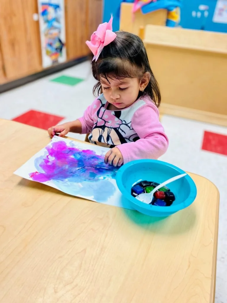 A young girl with a pink bow in her hair is painting with watercolors on a piece of paper at a wooden table.
