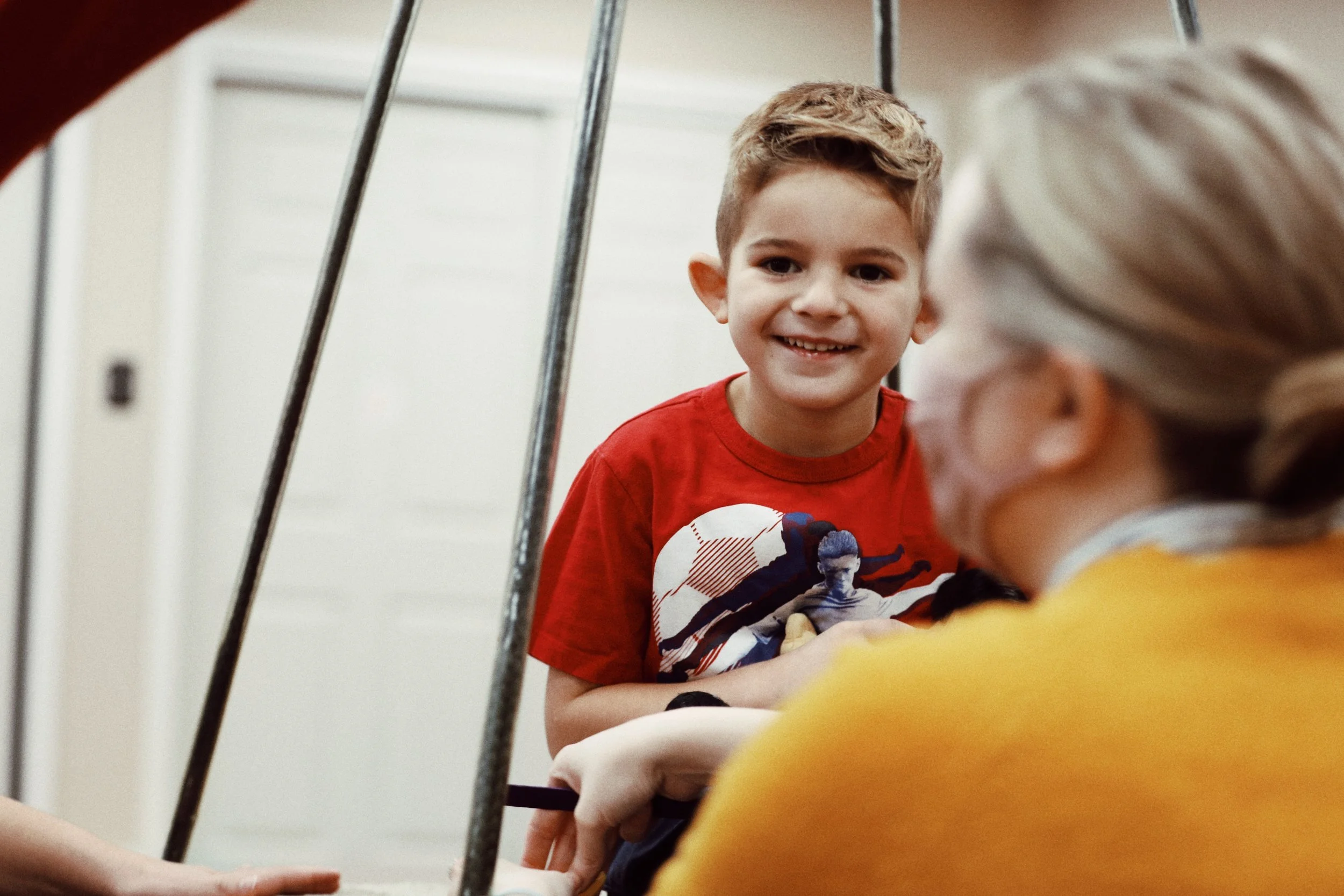 A young boy with brown hair and a red T-shirt smiling at an elderly woman with white hair, who is partially visible and wearing a yellow top, in a home setting.