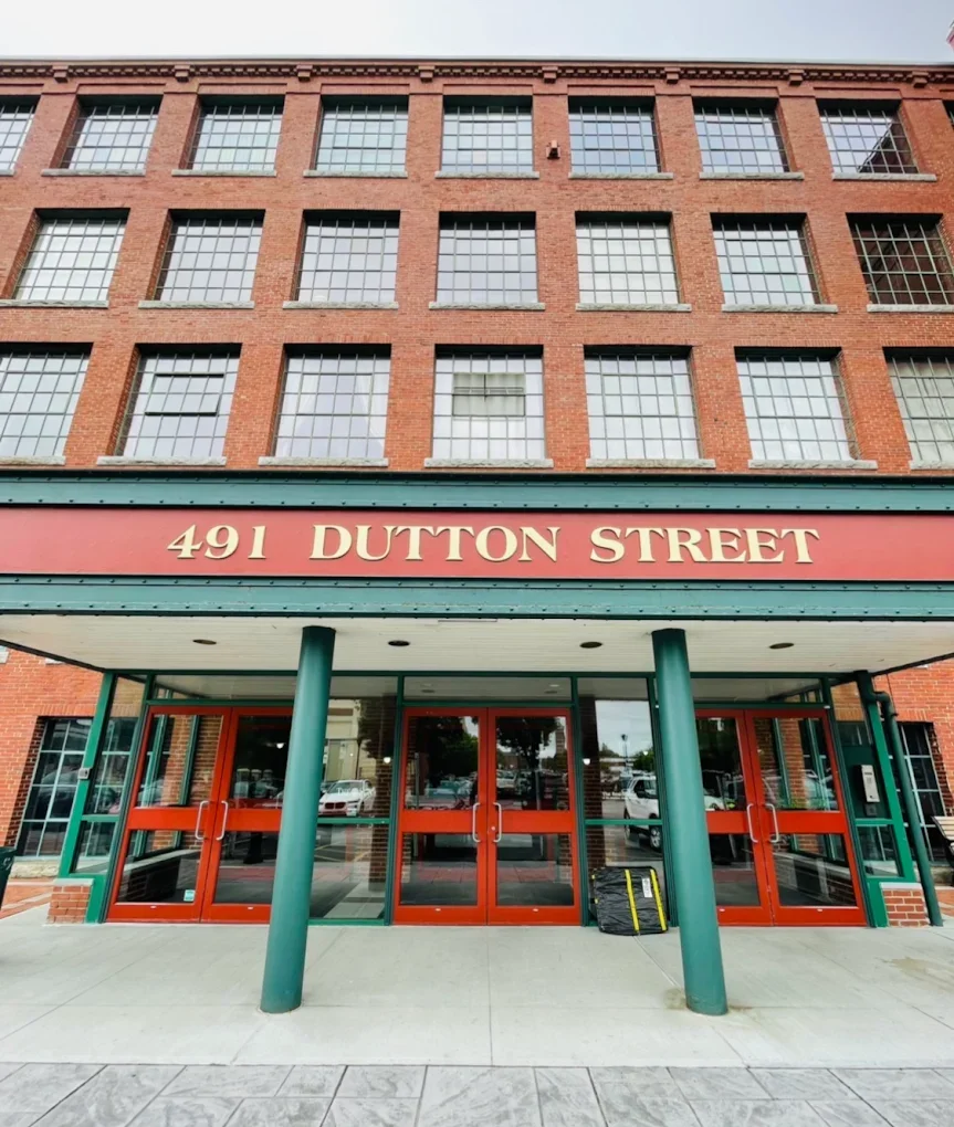 Building facade with red brick exterior and multiple large windows, entrance with glass doors, green and red color scheme, and a sign displaying "491 Dutton Street."