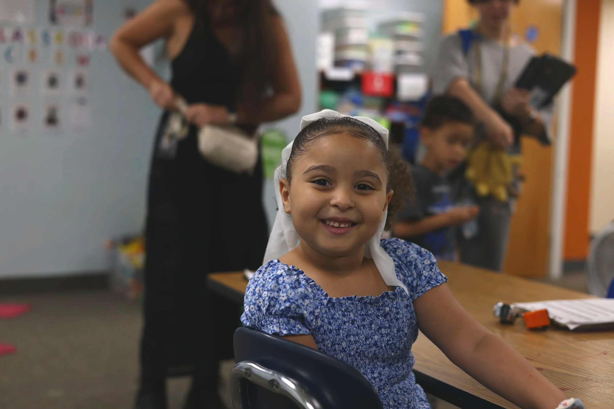 A young girl wearing a blue dress with a white bow in her hair, smiling at the camera, sitting at a table in a classroom. In the background, there are two adults and another child, with classroom shelves and colorful posters.