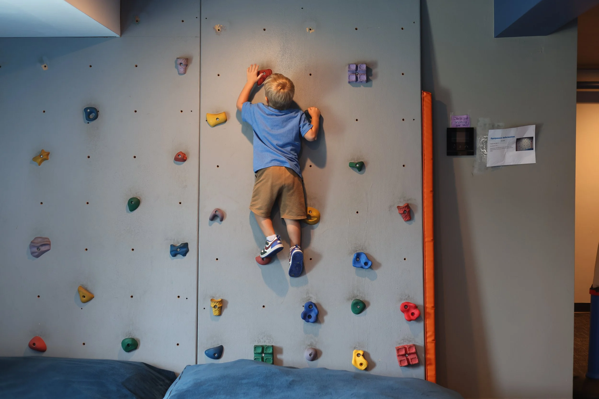 A young boy wearing a blue t-shirt and khaki shorts climbing an indoor rock climbing wall with various colorful holds.