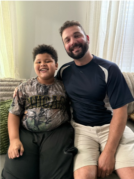 A smiling boy with curly hair sitting next to a smiling man with a beard on a beige couch in a well-lit living room.