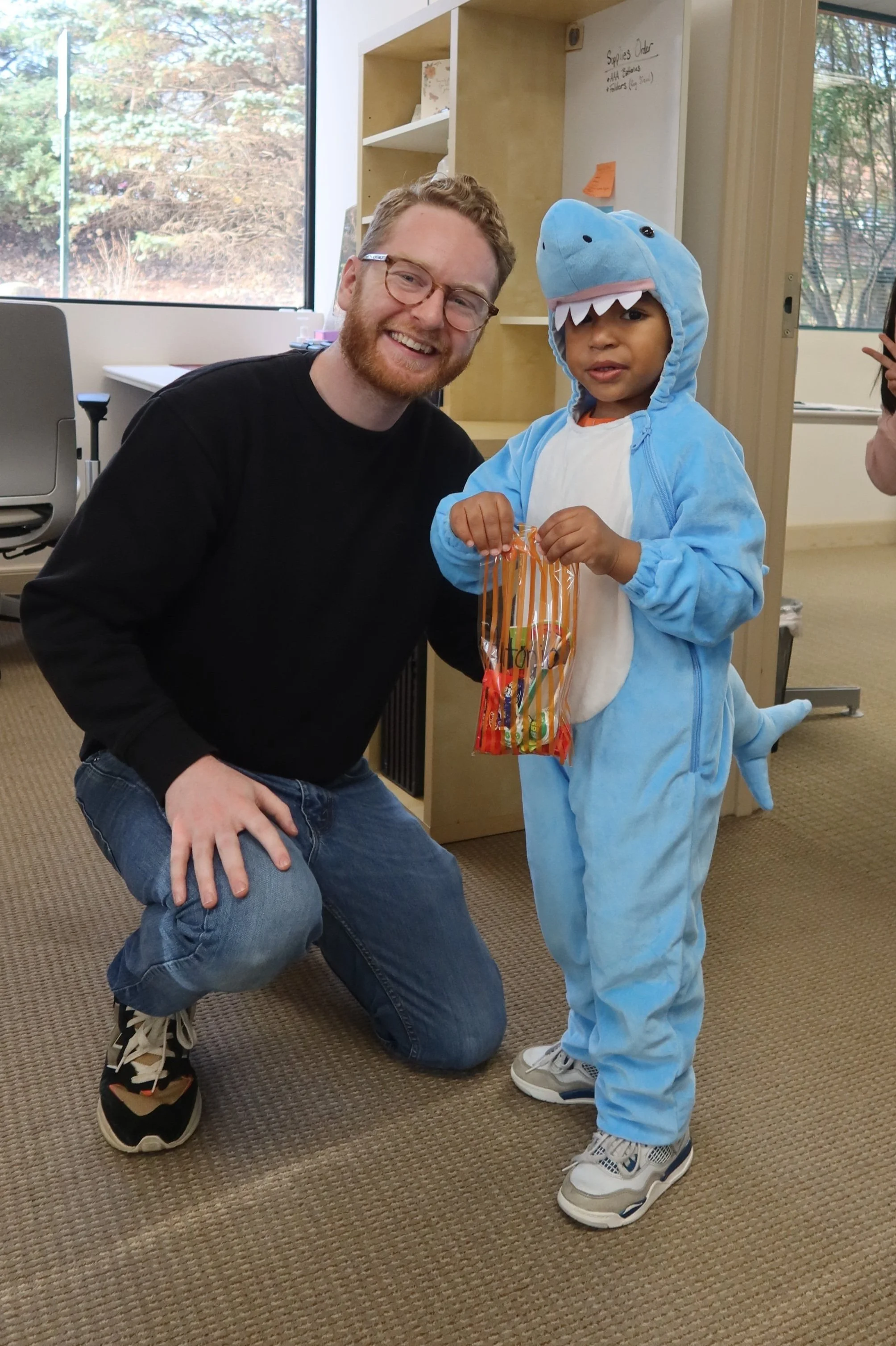 A man kneeling next to a young boy dressed in a blue shark costume, holding a plastic bag of candy, inside an office or classroom.
