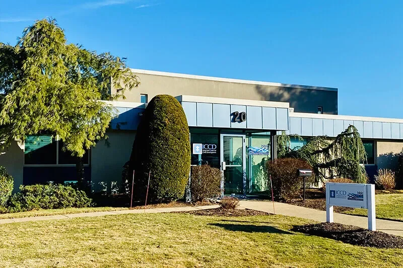 Exterior of an office building with large bushes and trees, a glass door entrance, and a sign that reads 'ICCD' in a small town in sunny weather.
