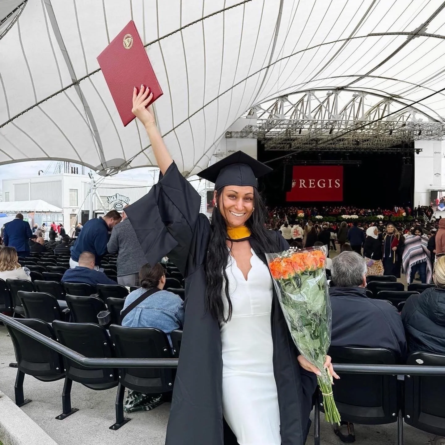 A graduate woman in a cap and gown holding a bouquet of flowers and celebrating at her graduation ceremony with a stage and seated audience in the background.