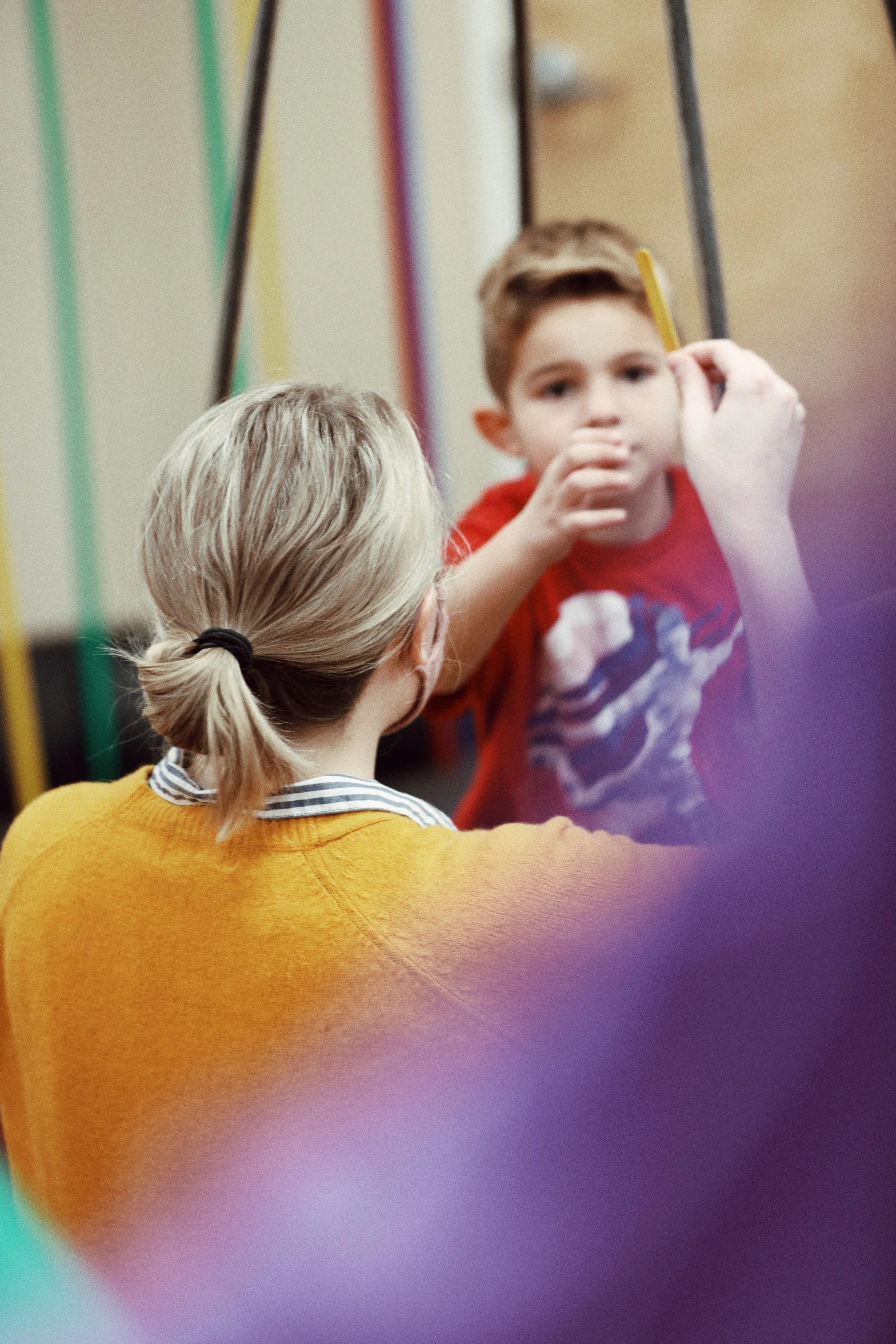 An adult woman with blonde hair tied back, seen from behind, listens while a young boy with short brown hair in a red shirt speaks and gestures with his hand, holding a yellow pencil. She is providing occupational therapy in a center-based setting.