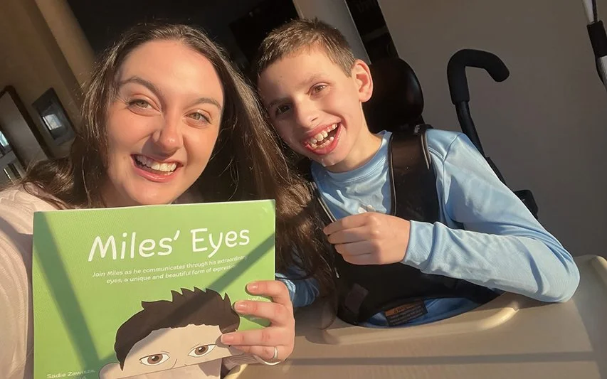 A woman and a young boy are sitting together, smiling for the camera. The boy is in a specialized chair with head and back support. The woman is holding a green book titled 'Miles' Eyes' with an illustration of a person with glasses on the cover.