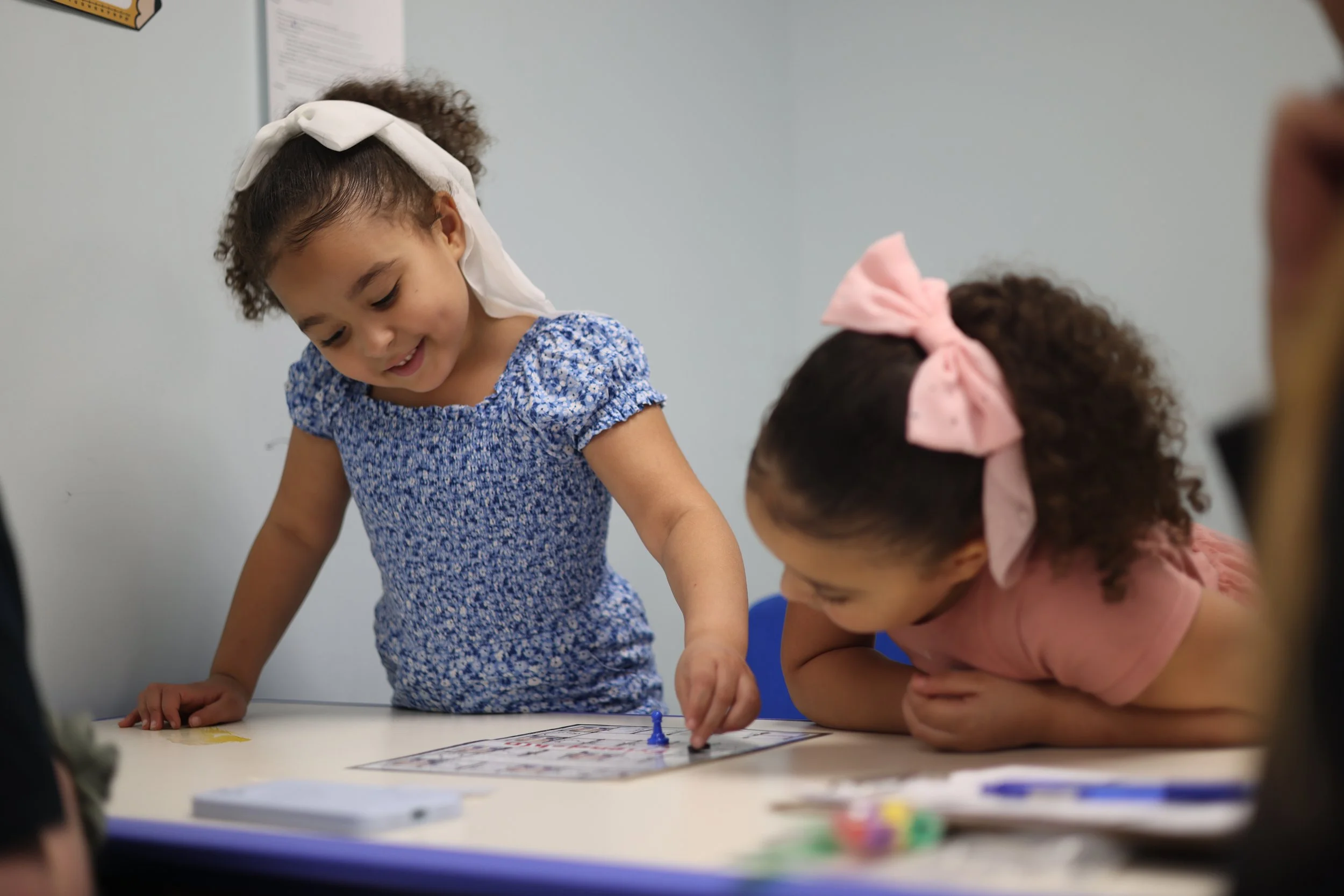 Two young girls with curly hair, one wearing a white hair bow and the other a pink hair bow, are playing a board game together at a table in a classroom.