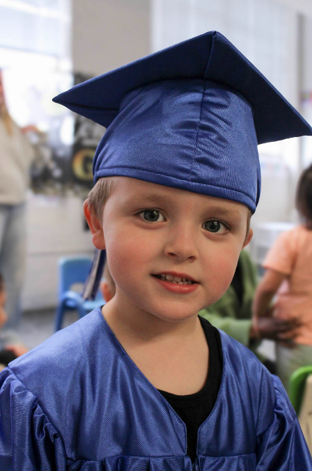 A young boy wearing a blue graduation cap and gown, smiling at the camera.