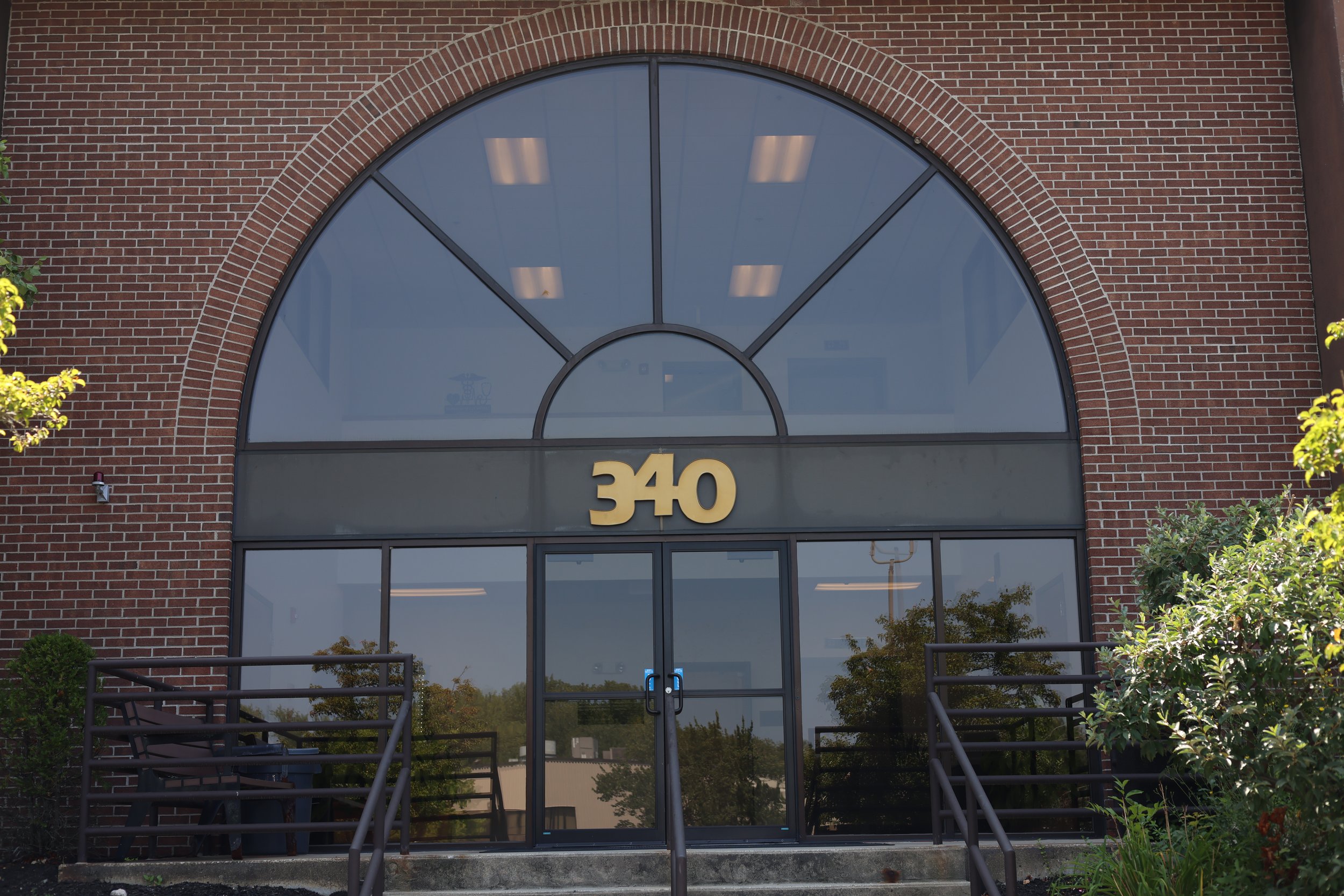The front entrance of a brick building with the number 340 in gold above the glass door, with an large arched window above, and railings on either side of the stairs leading to the door.