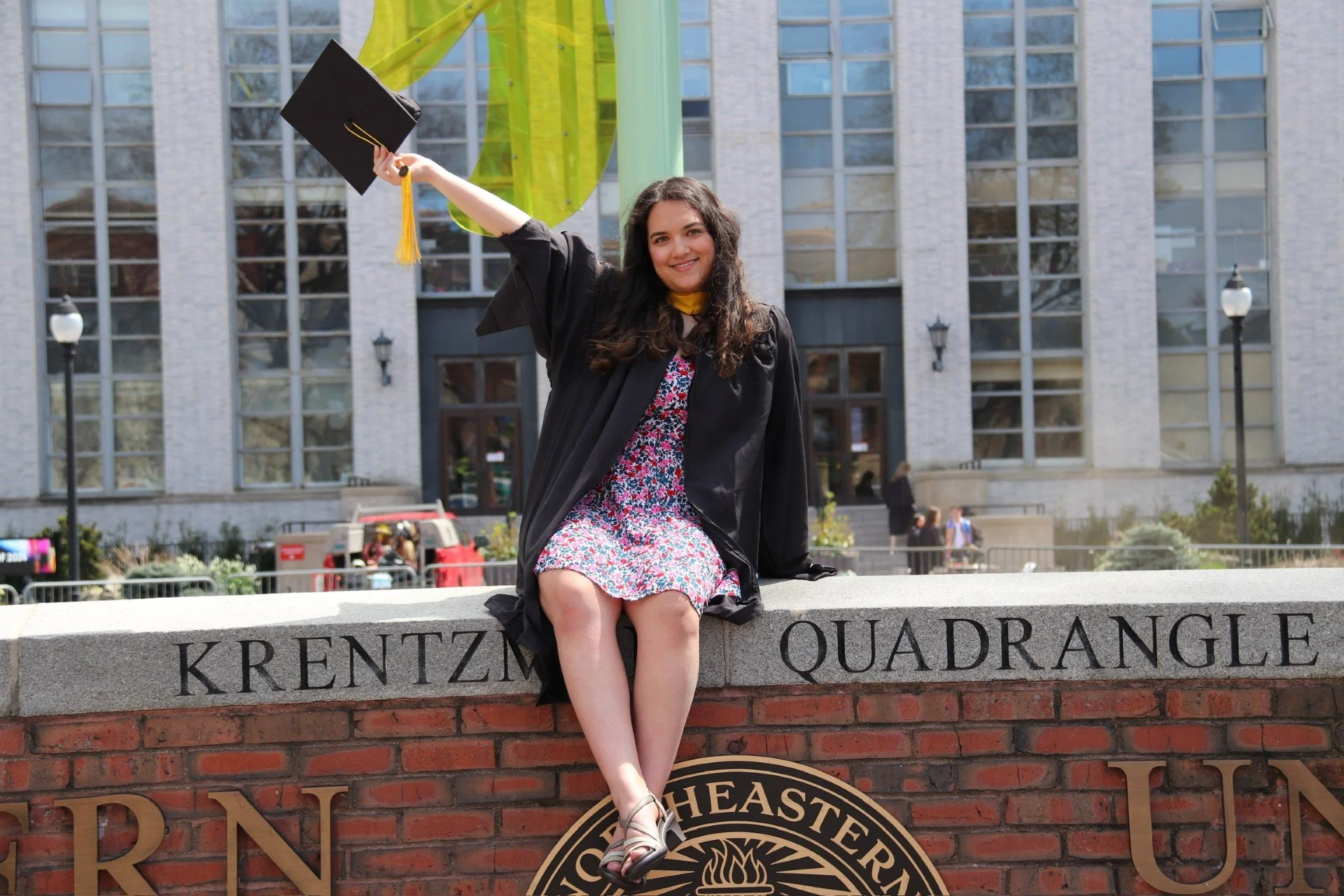 A young woman in a graduation cap and gown sitting on a bricks ledge with her legs hanging over the edge. She is smiling and holding her cap high in the air in front of a university building with large windows.