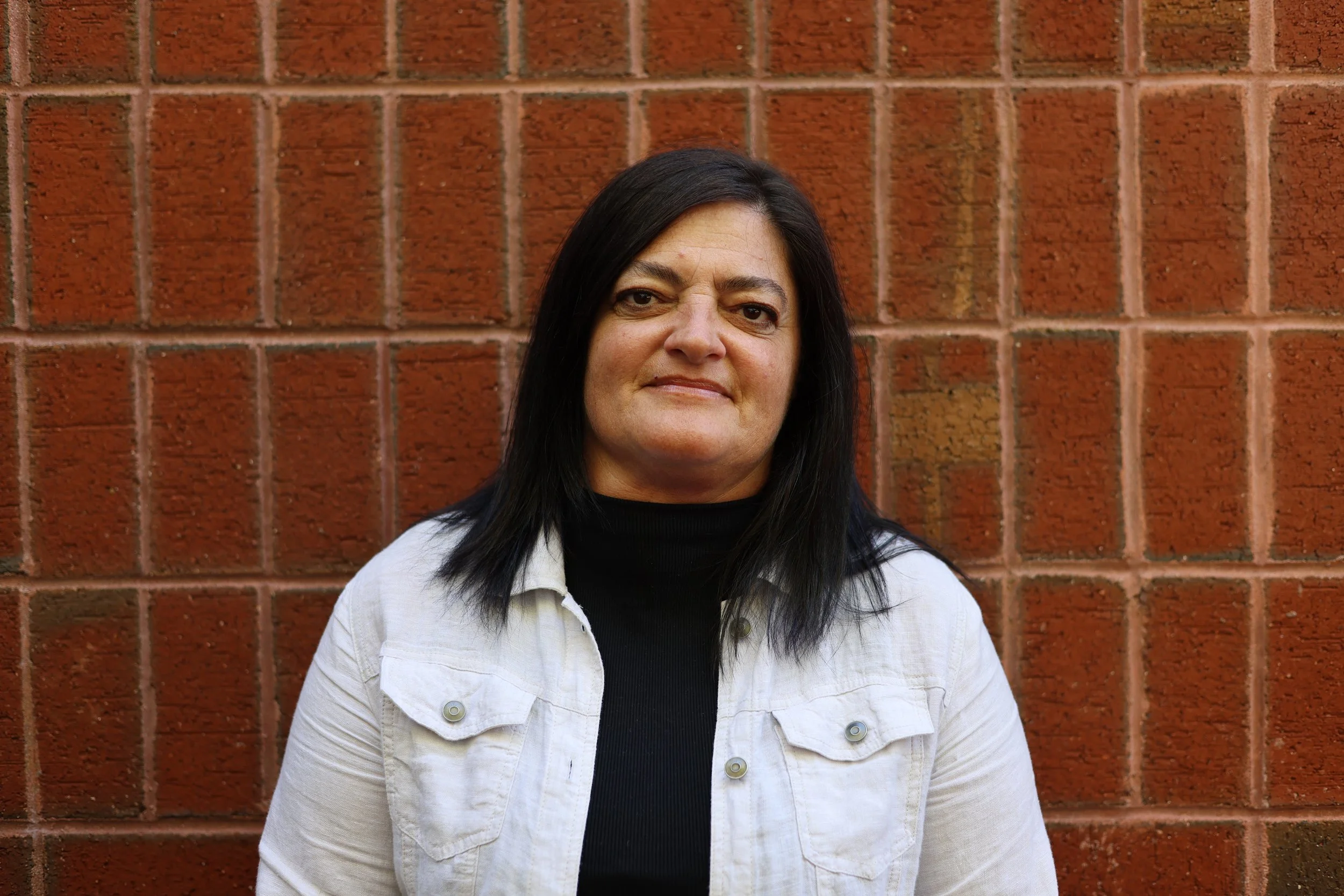 A woman with black hair and light skin standing against a red brick wall, wearing a white jacket over a black top, looking directly at the camera with a slight smile.