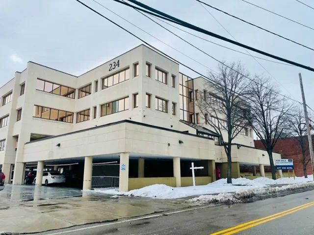 White multi-story apartment building with parking in front, snow on the ground, leafless trees, and a tree-lined sidewalk.