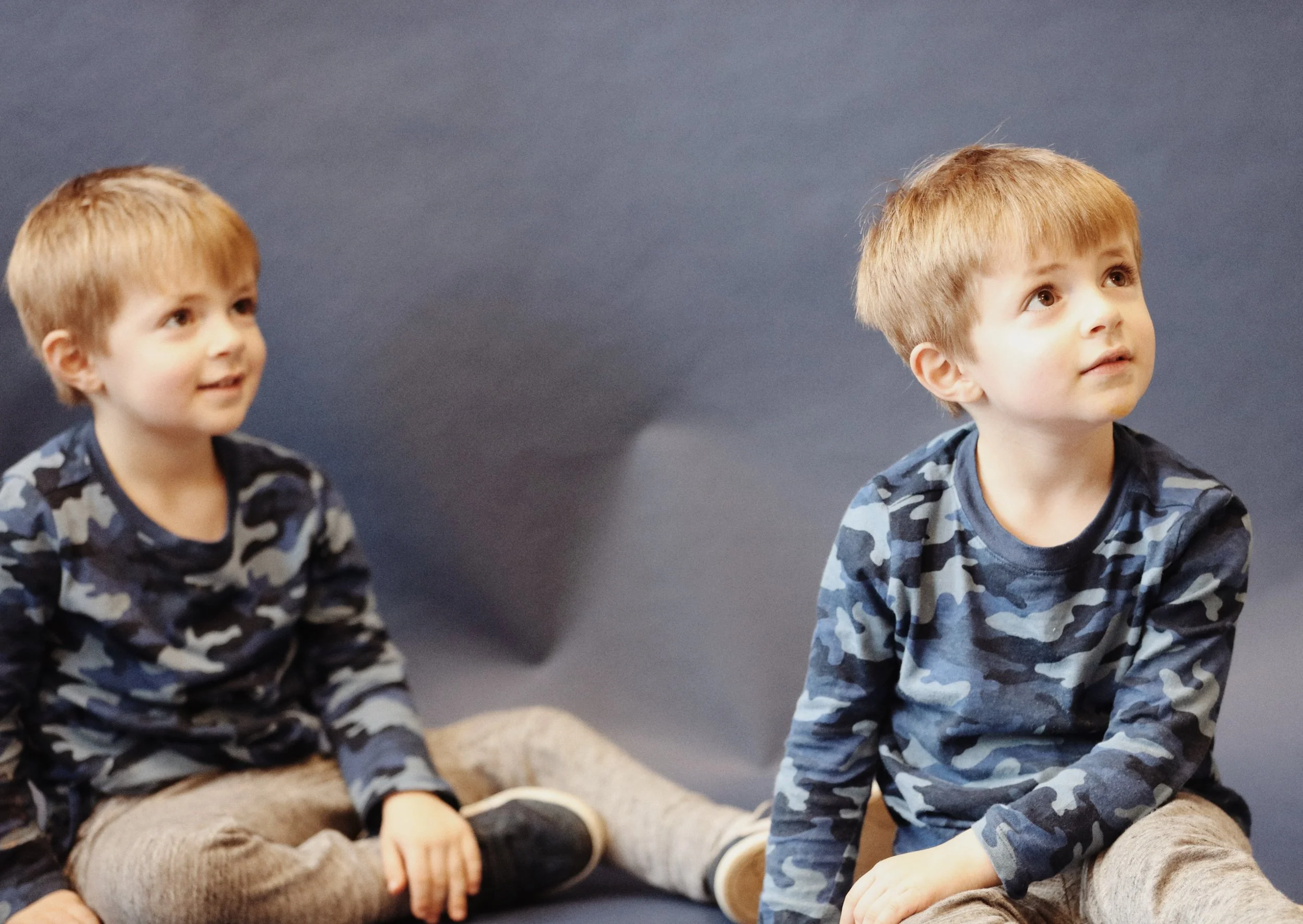 Two young boys with blond hair, dressed in matching blue camouflage shirts and beige pants, sitting on a dark blue surface against a dark blue background. One boy is sitting cross-legged, smiling and looking to his left, while the other boy is sitting with his legs stretched out, looking up and to his right.