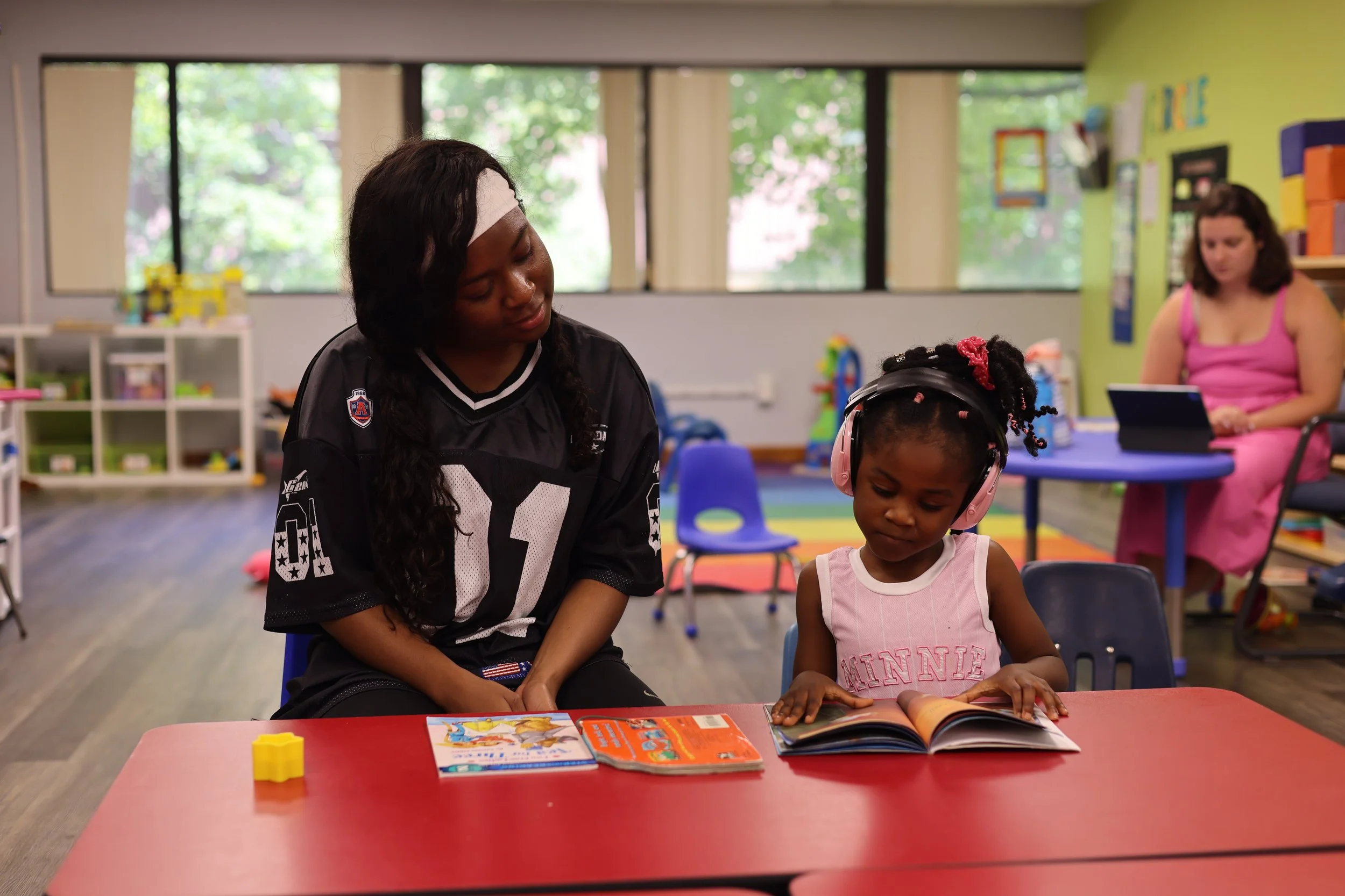 A woman and a child sitting at a table in a classroom, reading books. The woman is wearing a black sports jersey, and the child is wearing a pink tank top and pink headphones. In the background, another woman is sitting at a table using a tablet.