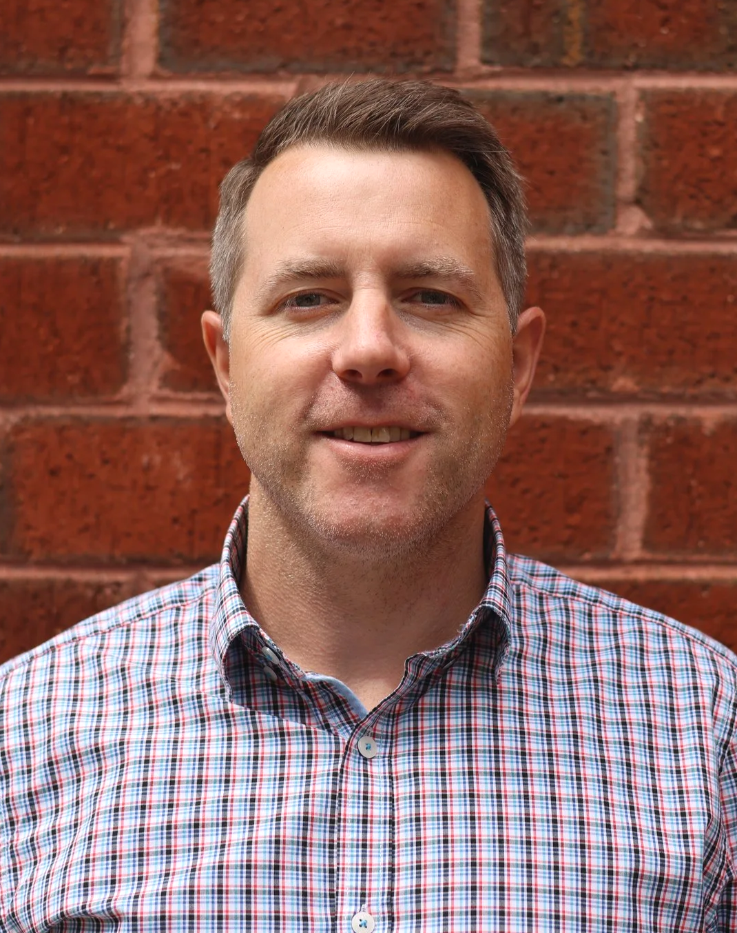 A man with short brown hair, wearing a red, blue, and white plaid shirt, standing in front of a red brick wall.